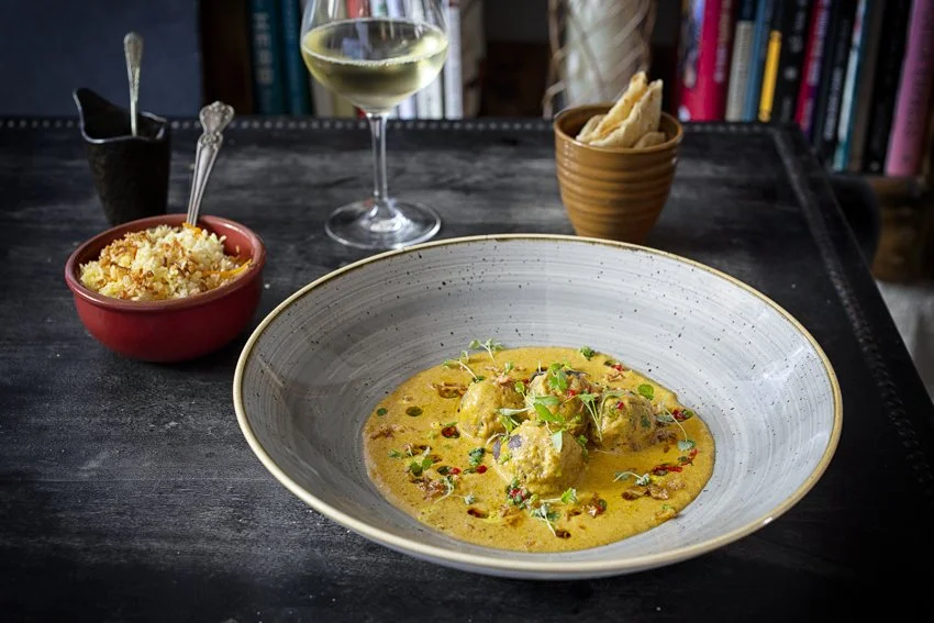 A bowl of yellow curry with chicken and garnished with microgreens, on a dark wooden table with a glass of white wine, a bowl of rice, and a small bowl of breadsticks in the background.