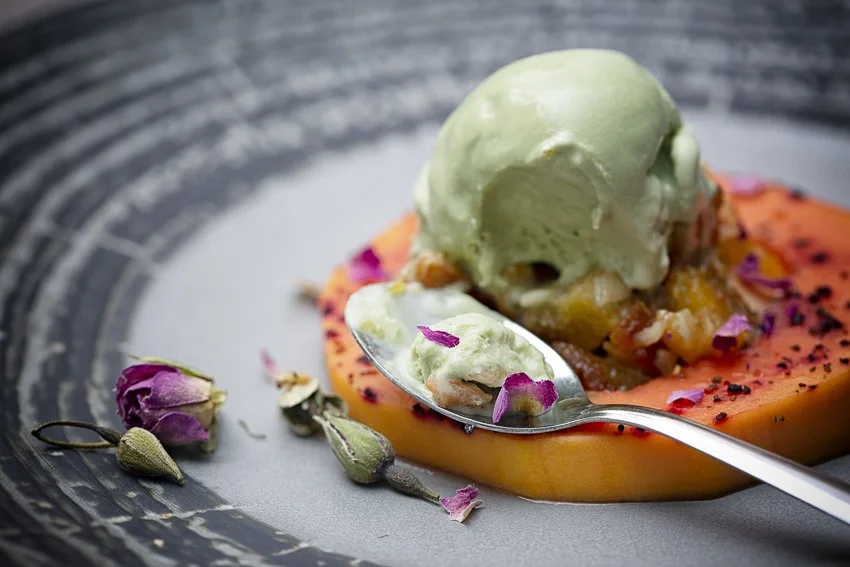 Dessert with a scoop of green ice cream on top of a slice of orange-colored cake, garnished with dried flower petals and edible flowers on a black textured plate.