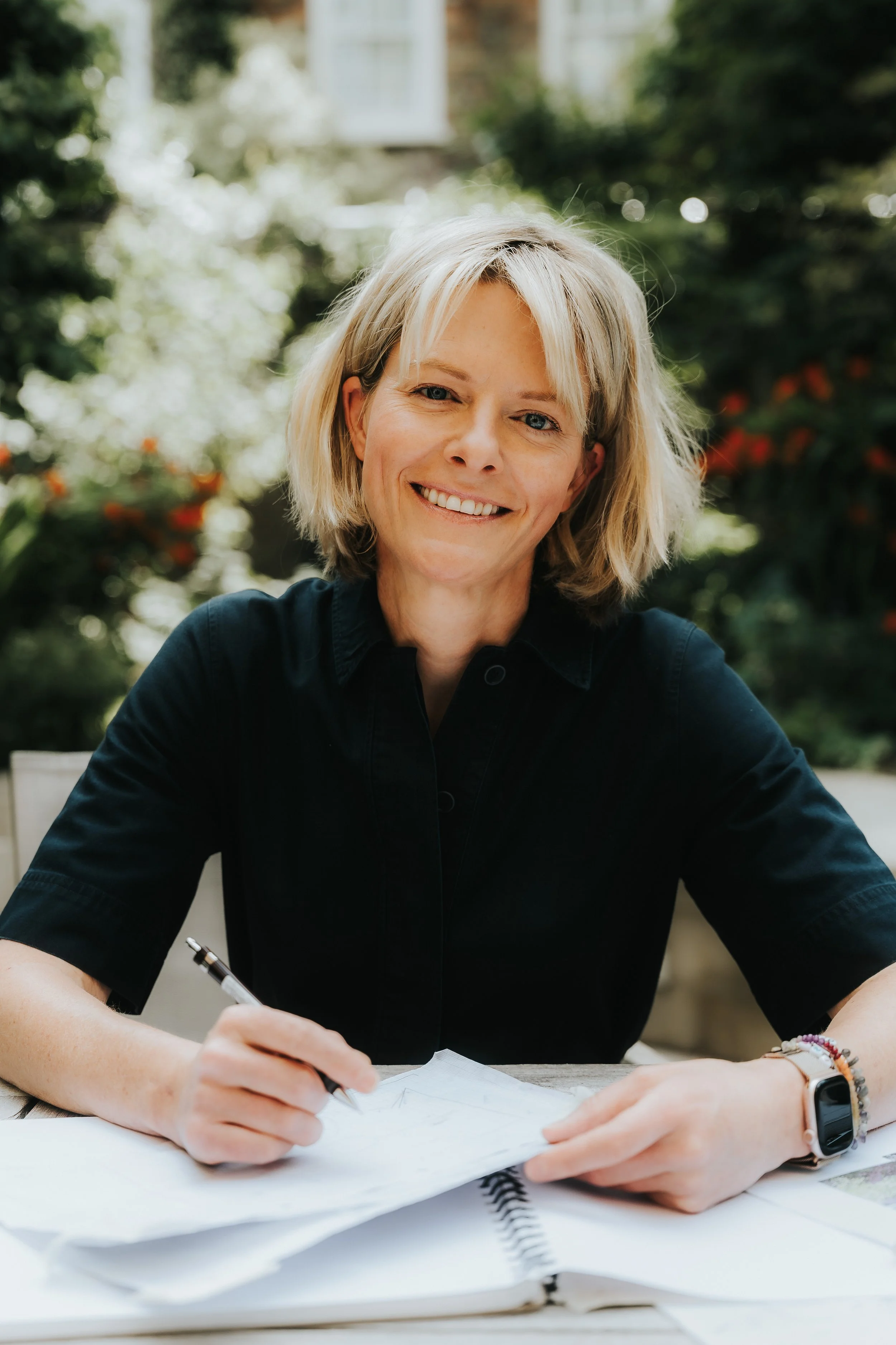 A woman with blonde hair smiling and writing with a pen at a desk outdoors, with greenery and a partial view of a house in the background.