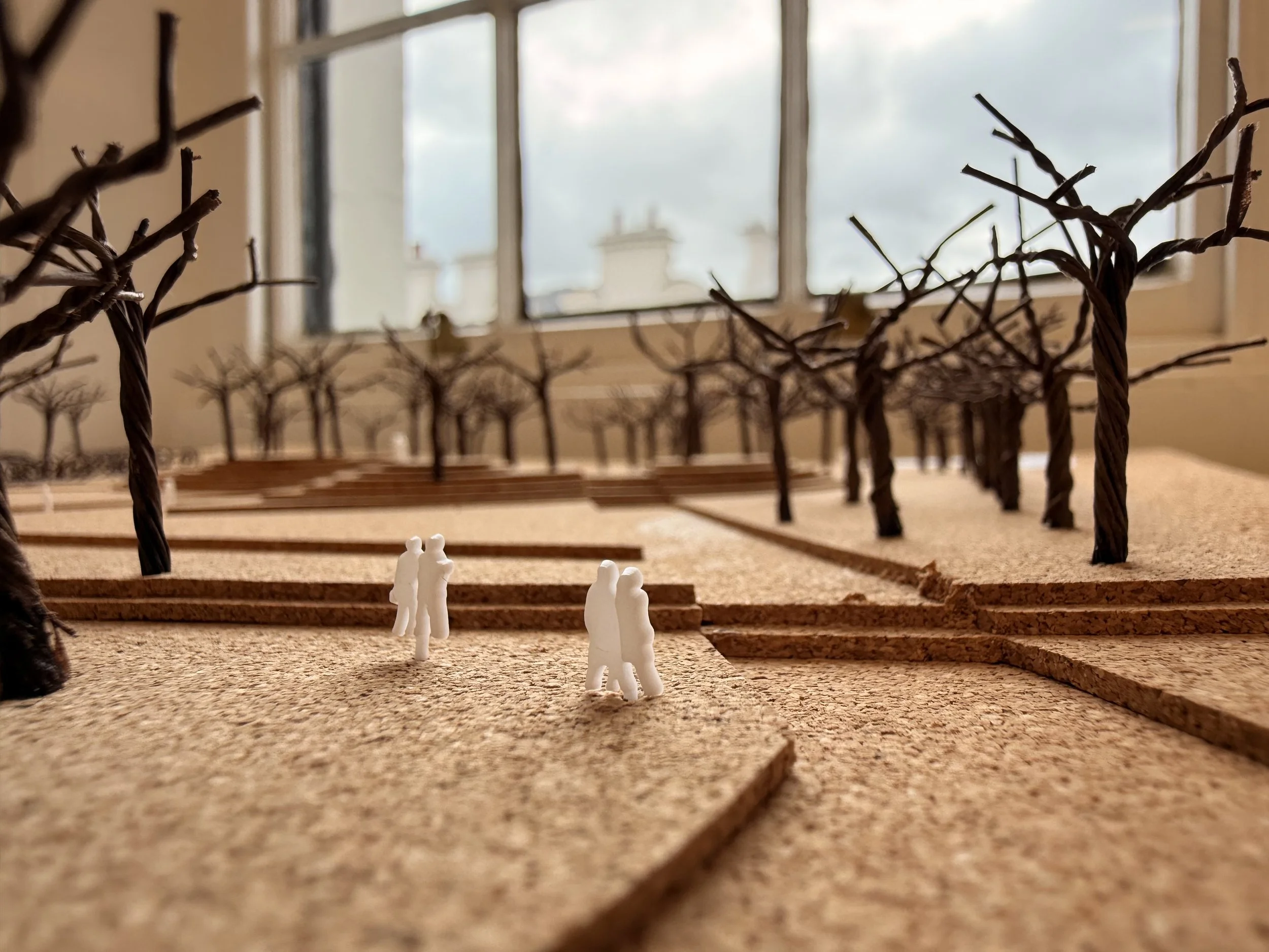 Miniature white figurines of people walking on a textured surface, with a backdrop of leafless tree branch models and a window showing an outdoor scene with a cloudy sky and building rooftops.