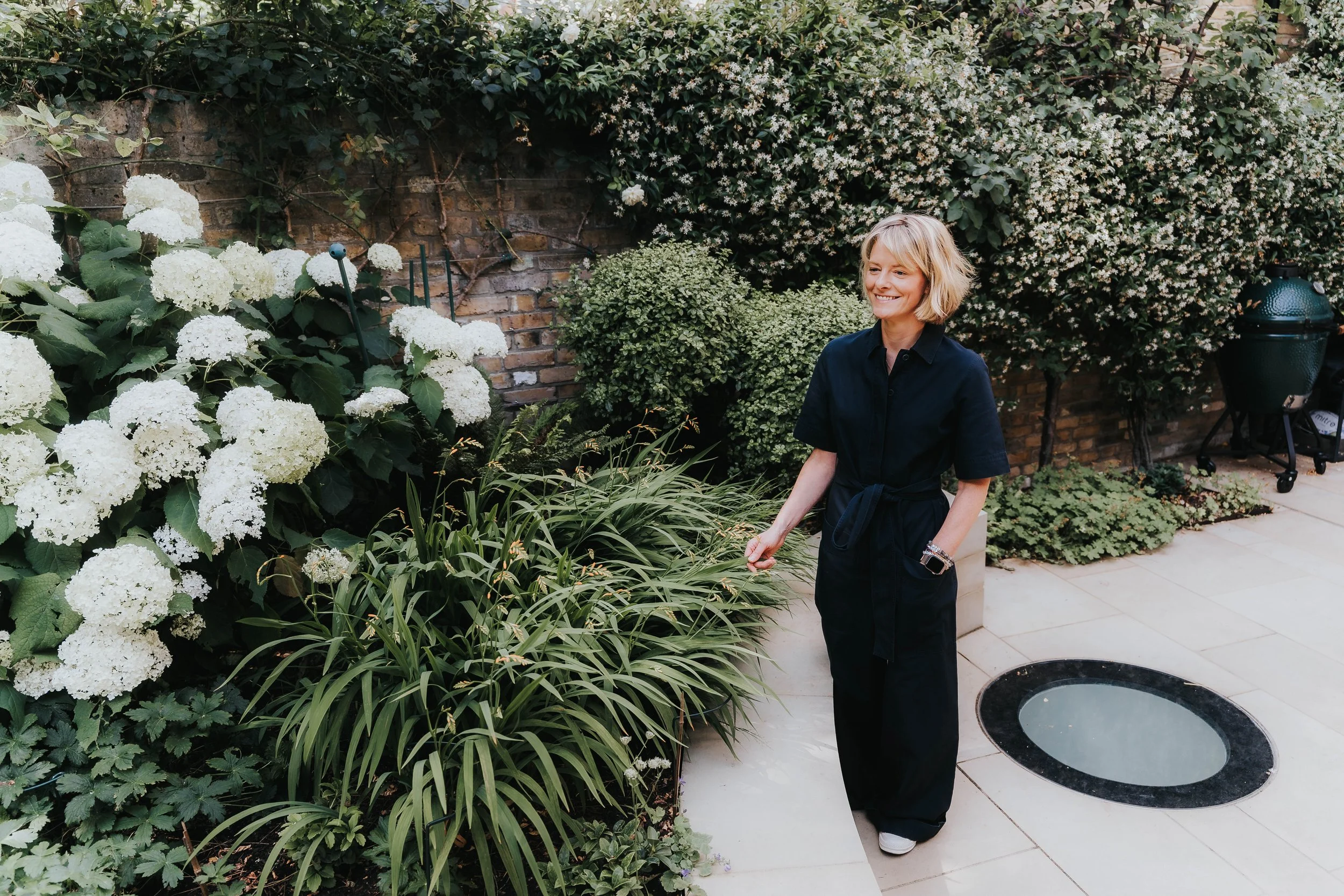 A smiling woman with blonde hair in black clothing and white shoes walking in a lush garden with white flowers, green plants, and a brick wall background.