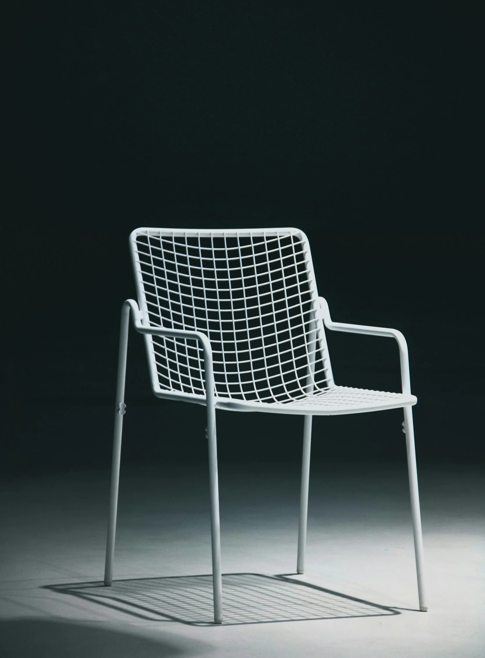 A white mesh chair with armrests casting a shadow on a light-colored floor against a dark background.
