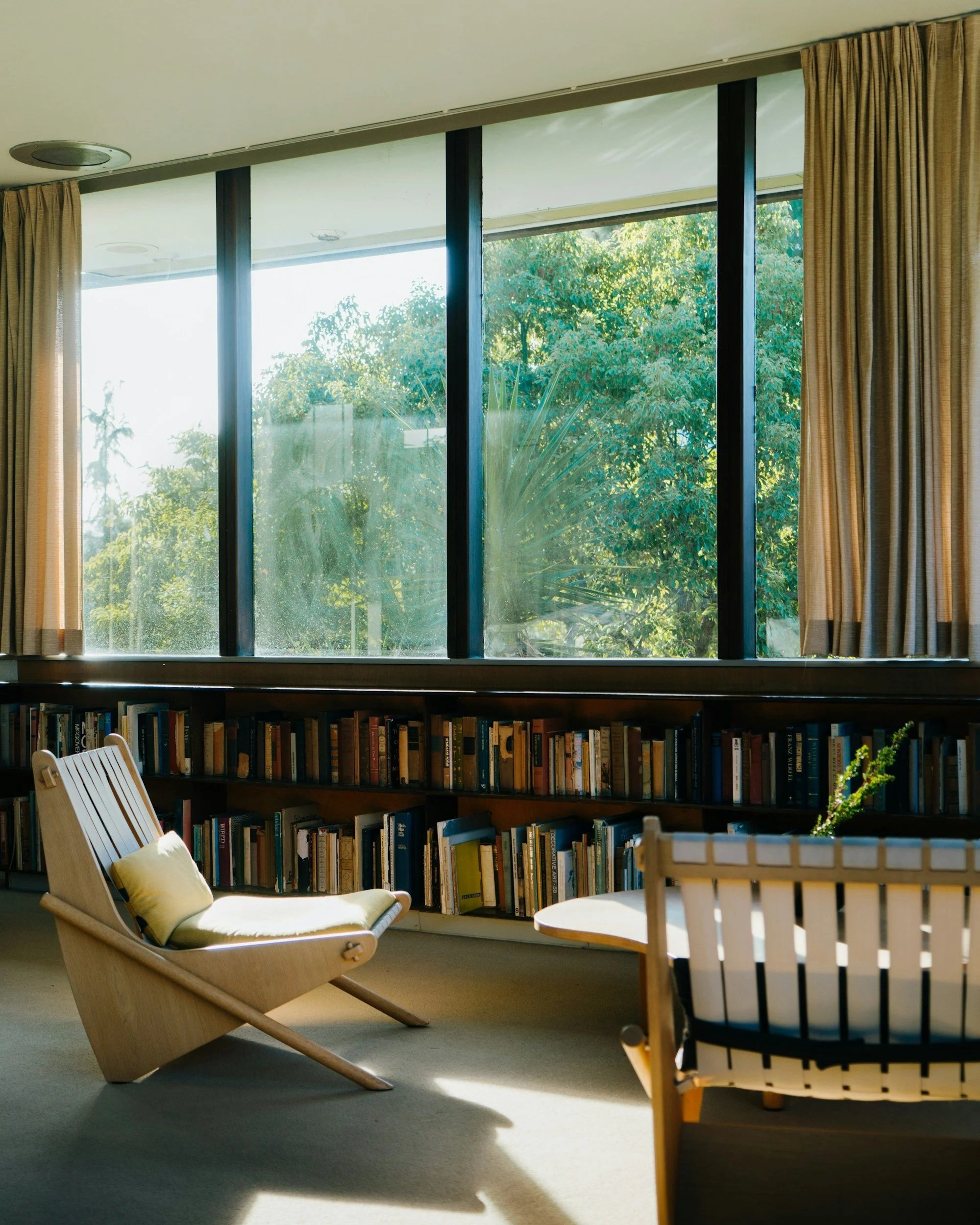Living room with large window showing green trees outside, containing a beige lounge chair with a yellow pillow, a round wooden table, and a black bookshelf filled with books.