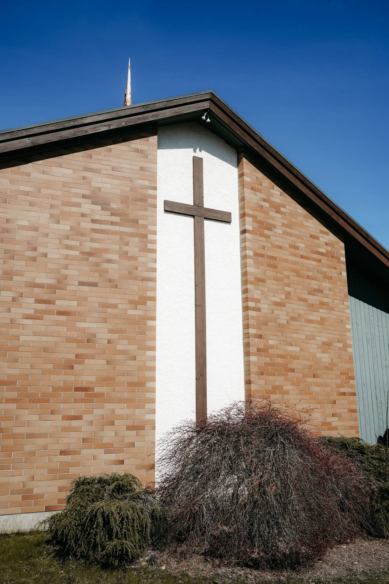 A brick church building with a white section in the middle featuring a wooden cross. There are dark bushes and grass in front, under a clear blue sky.