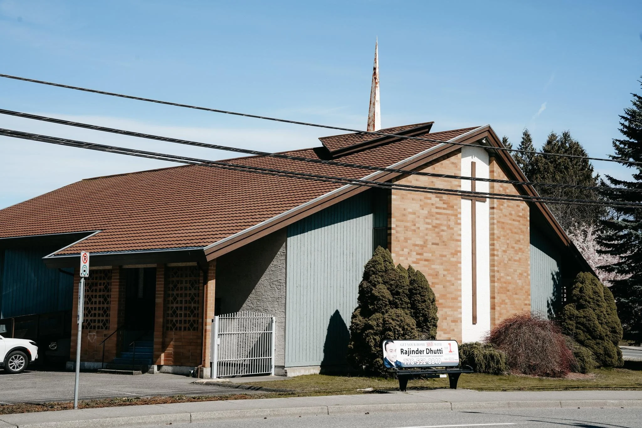 A residential building with a brown tiled roof, brick and light blue siding, and a small front yard with trees and bushes. There is a 'For Sale' sign featuring Rajinder Dhutti with his contact number in front of the house.
