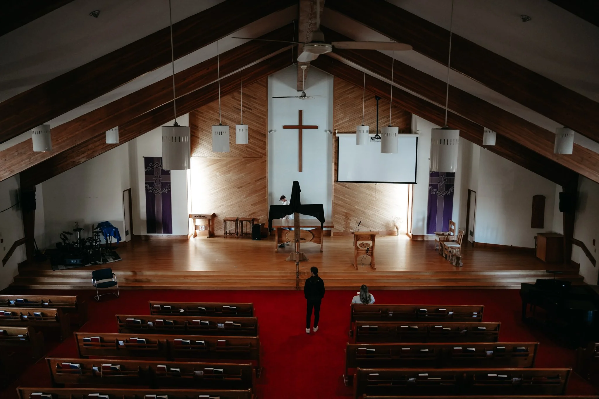 Inside view of a church with wooden ceiling beams, hanging white pendant lights, a cross on the wall, a large projection screen, a person standing near the altar, and empty pews with a few people.