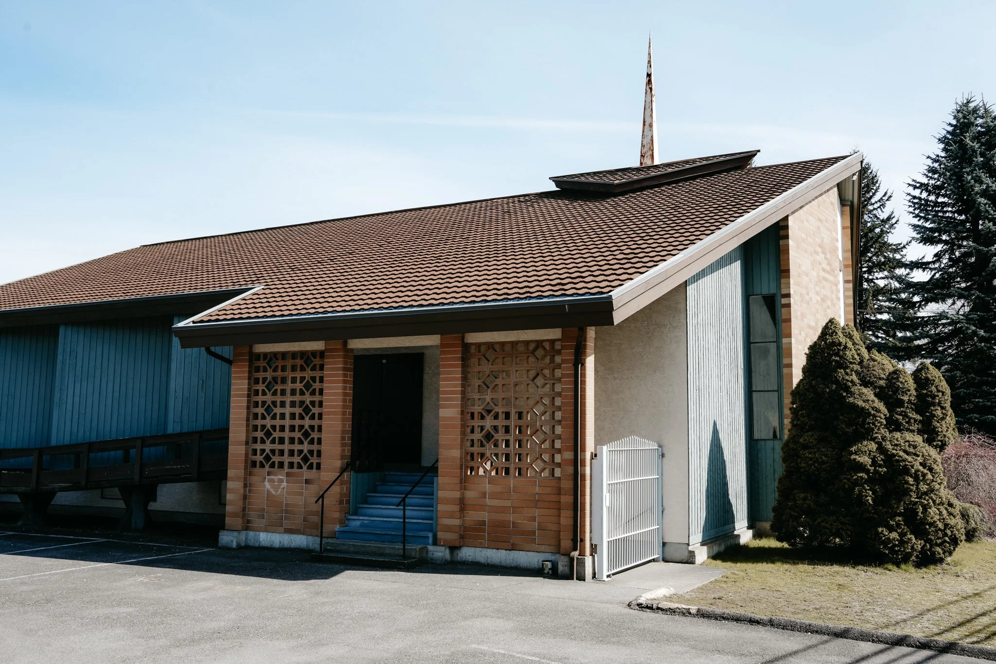 A small building with a sloped tile roof, a balcony made of wood, a staircase, brick walls, and a white fence. Tall trees are in the background under a clear blue sky.