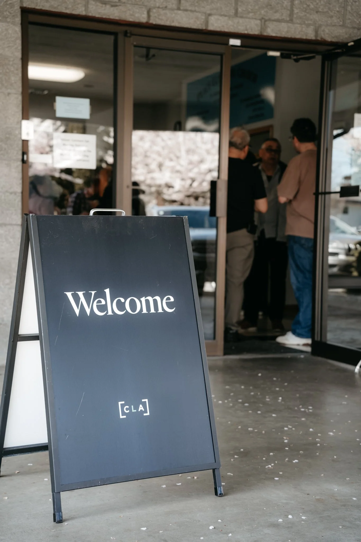 A black sandwich board sign with white text reading 'Welcome' and '[CLA]' standing on the sidewalk outside a building entrance. In the background, a glass door is open, revealing a group of people gathered inside, talking. The building has a brick exterior, and the ground is scattered with small white debris.