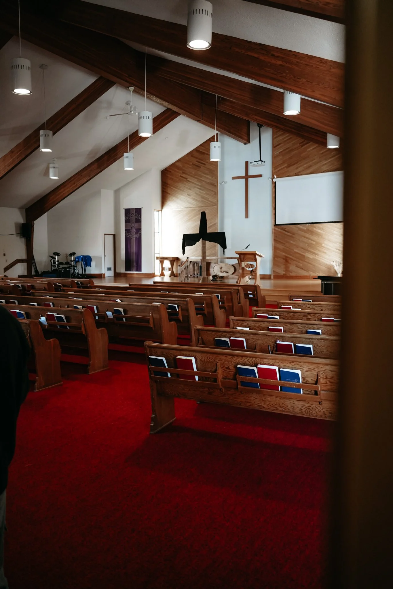 Interior of a church with wooden ceiling beams, rows of pews with hymnals, a cross on the wall, a pulpit, and a red carpeted aisle.