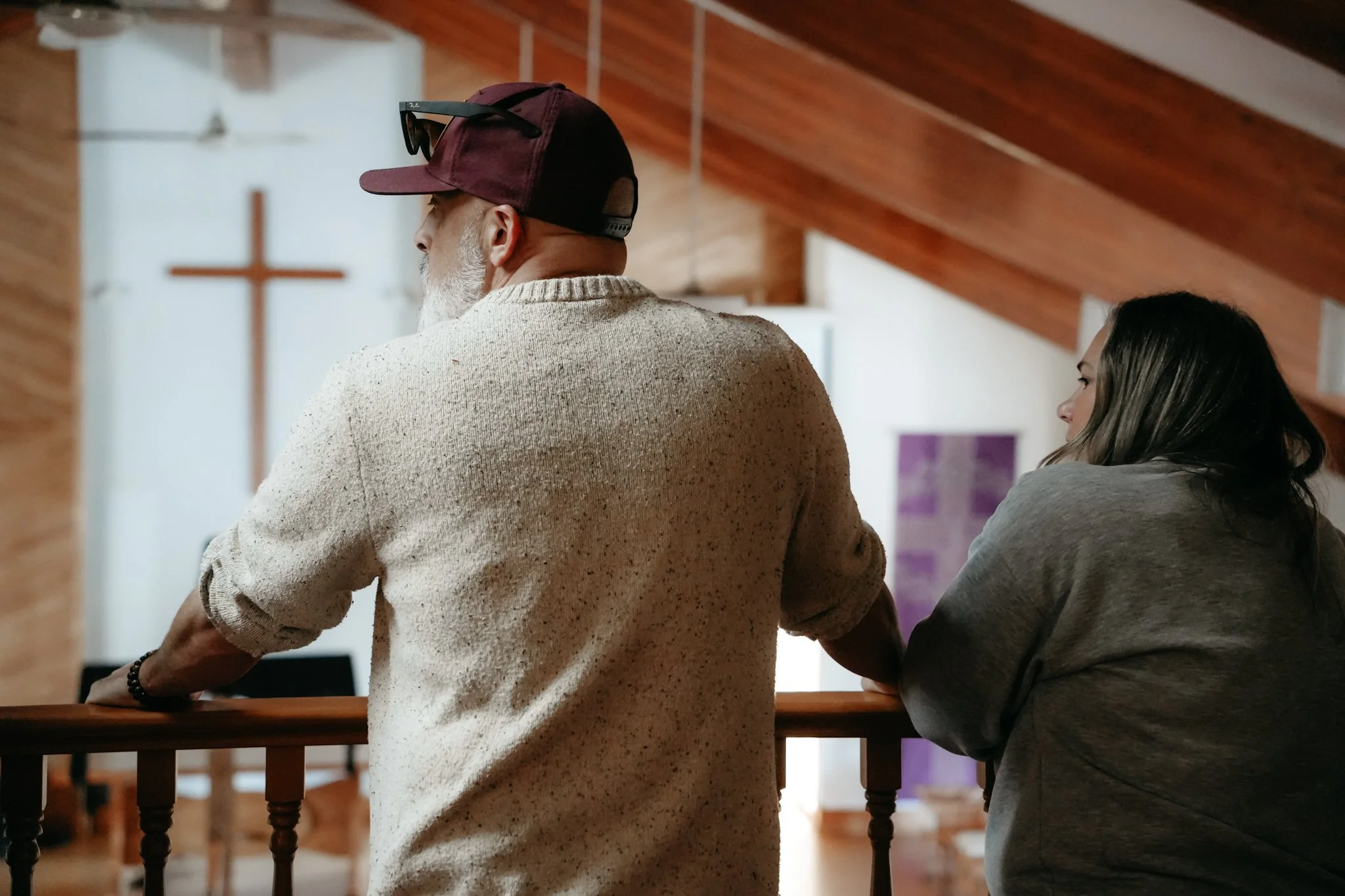 A man with a gray beard wearing a beige sweater, baseball cap, and sunglasses on his head, standing at a church railing. A woman with long dark hair, wearing a gray hoodie, stands next to him. The background shows a white wall with a red cross and a purple banner, wooden ceiling, and a ceiling fan.