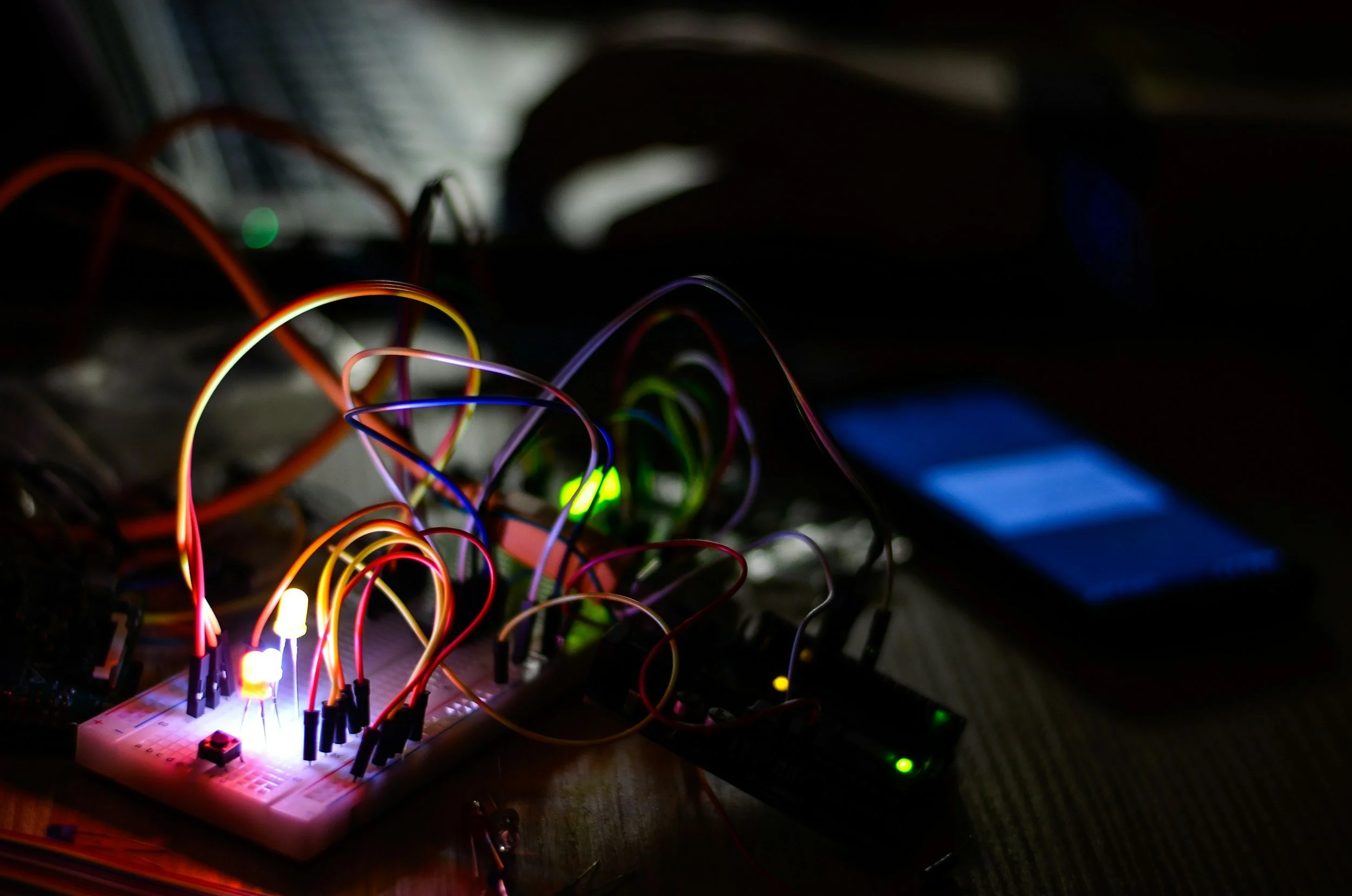 A close-up of a breadboard circuit with various colorful wires connected to LEDs and electronic components, illuminated by small LED lights in a dark environment.