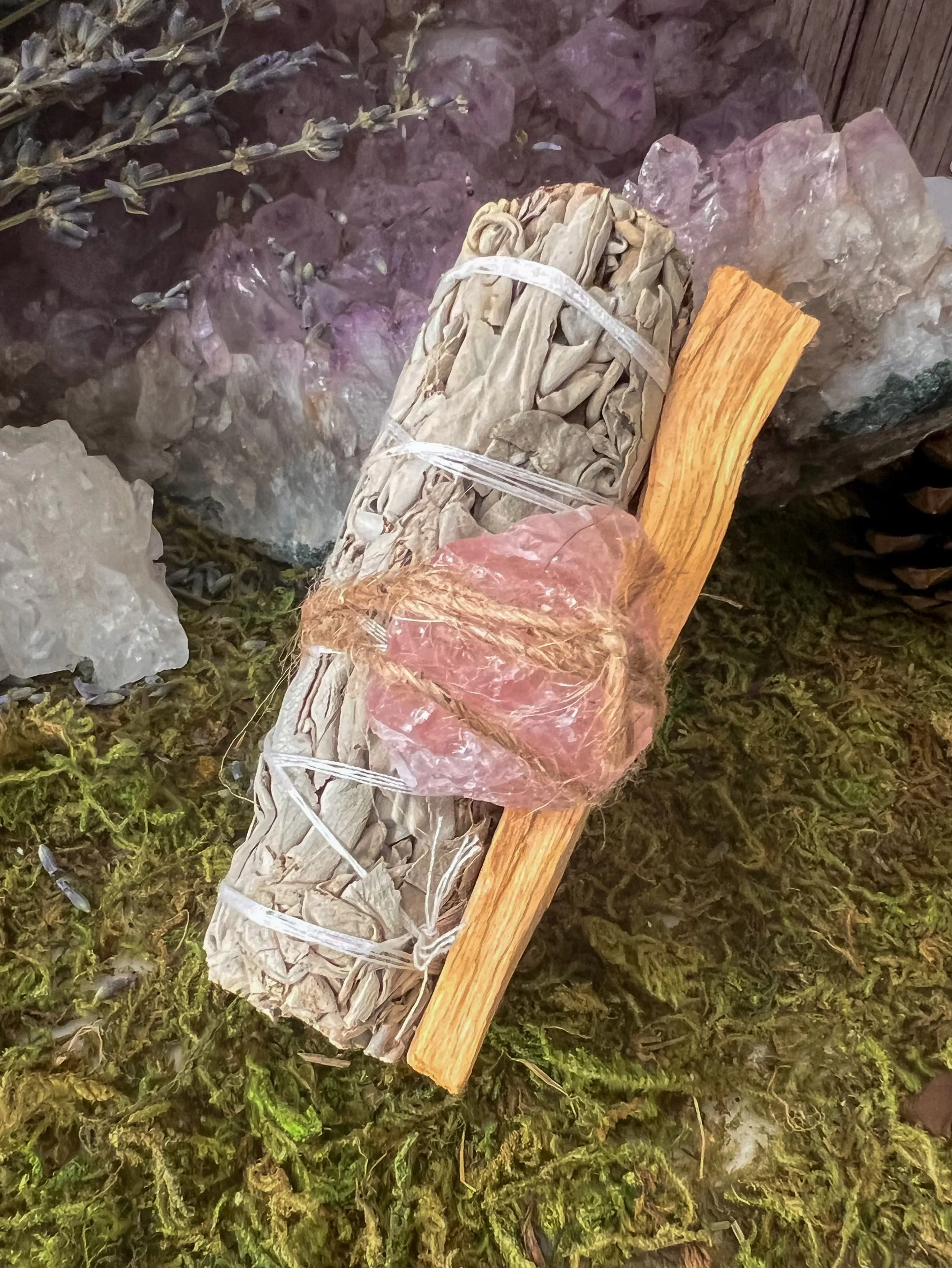 A bundle of sage and a piece of palo santo wood resting on green moss and rocks.