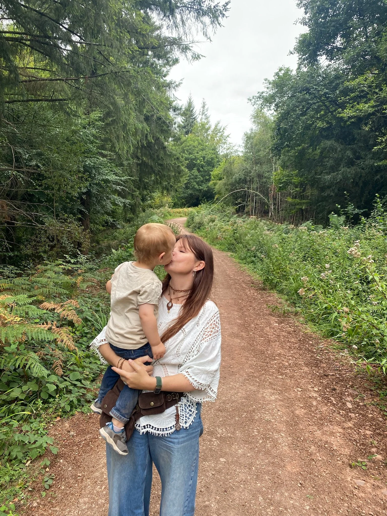 A woman with long brown hair wearing a white crochet top and blue jeans is holding a young boy with blond hair, a beige shirt, and blue jeans while kissing him on the lips on a dirt trail in a lush green forest.