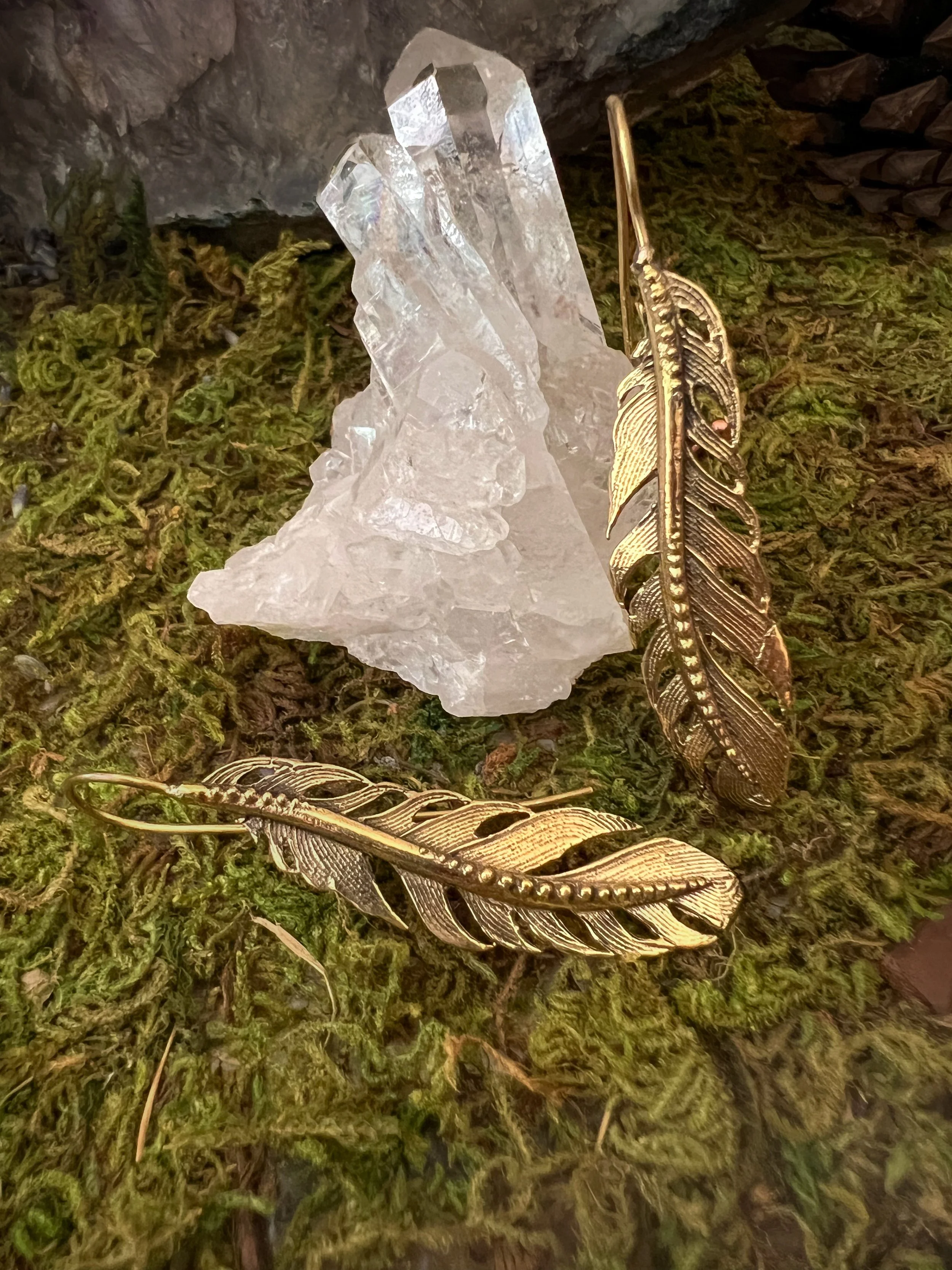 Close-up of a metallic, golden feather and clear quartz crystal on a mossy tree trunk.