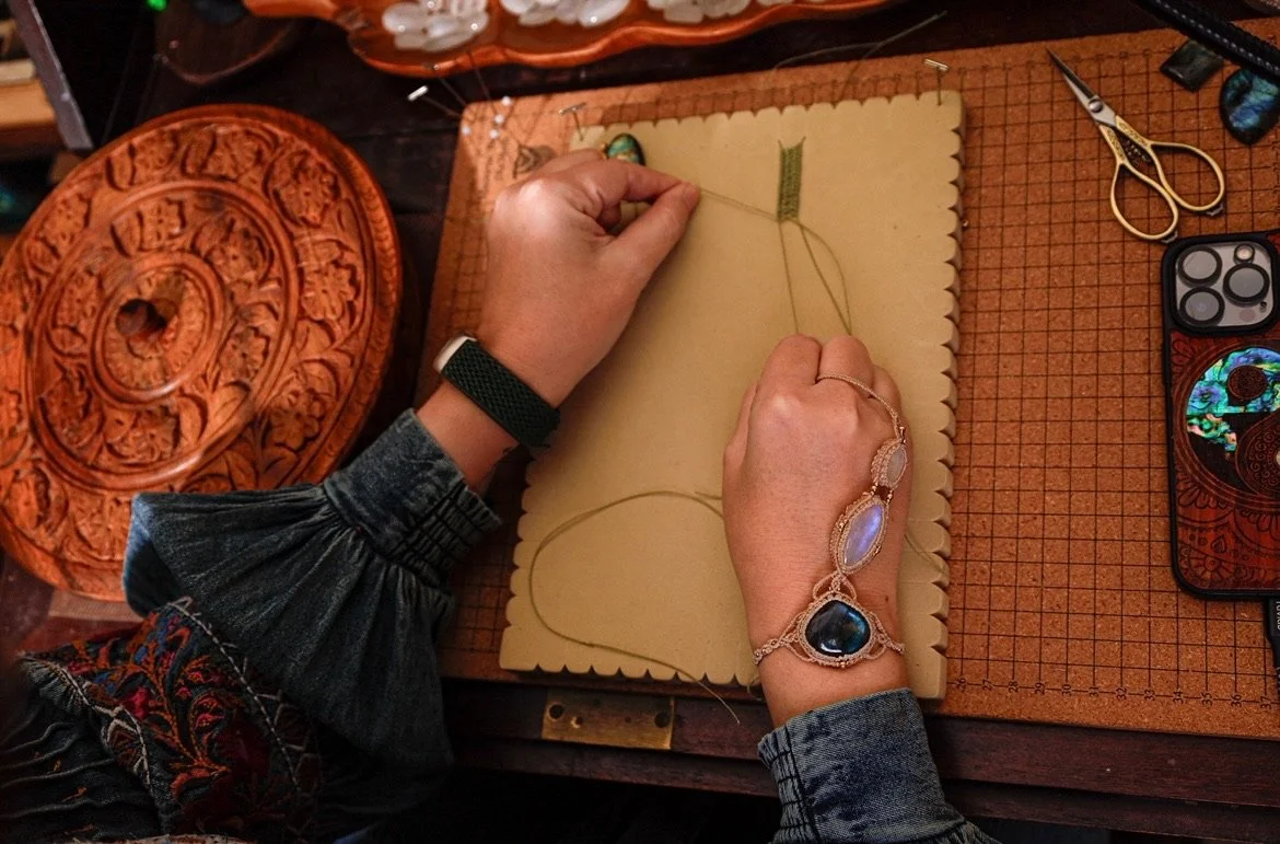 Person working on a needlework project, wrapping thread around a needle on a piece of fabric, with sewing tools like scissors nearby, on a wooden desk with decorative carved wooden objects.