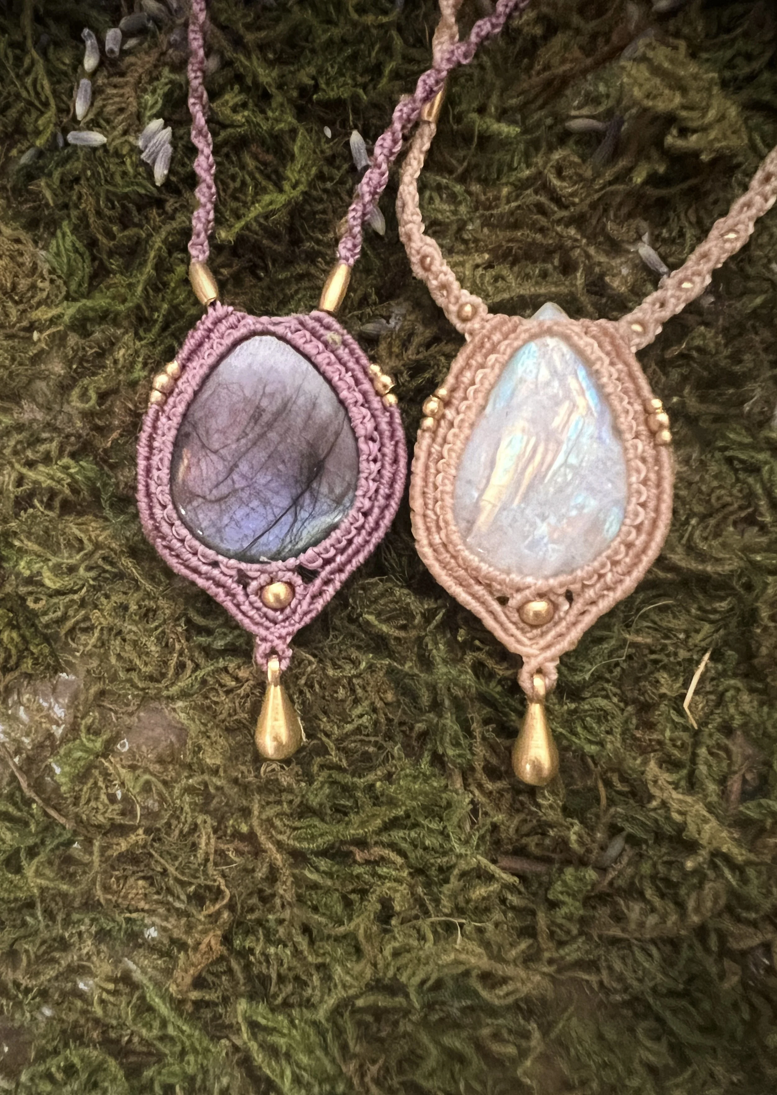 Two handcrafted necklaces with iridescent oval pendants, one with pink and purple cords and the other with beige cords, adorned with golden beads and teardrop charms, resting on a mossy surface.
