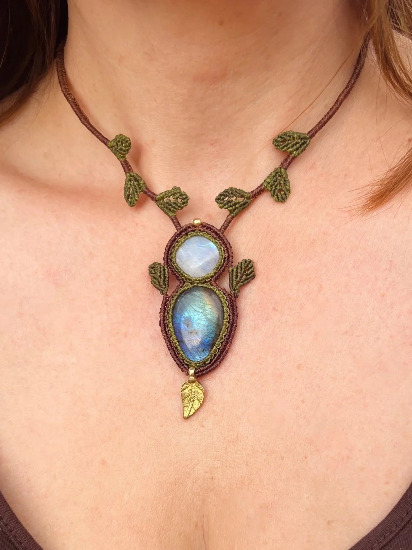 Close-up of a woman's neck wearing a elaborate necklace featuring two large iridescent stones, green leaf-shaped decorations, a golden leaf pendant, and brown cord.