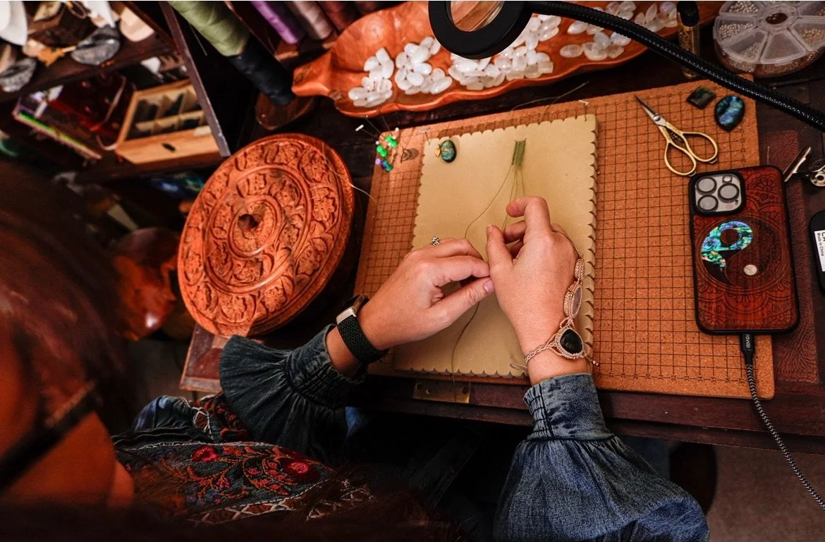 A person working on jewelry making at a cluttered workspace with tools, jewelry stones, and decorative items.