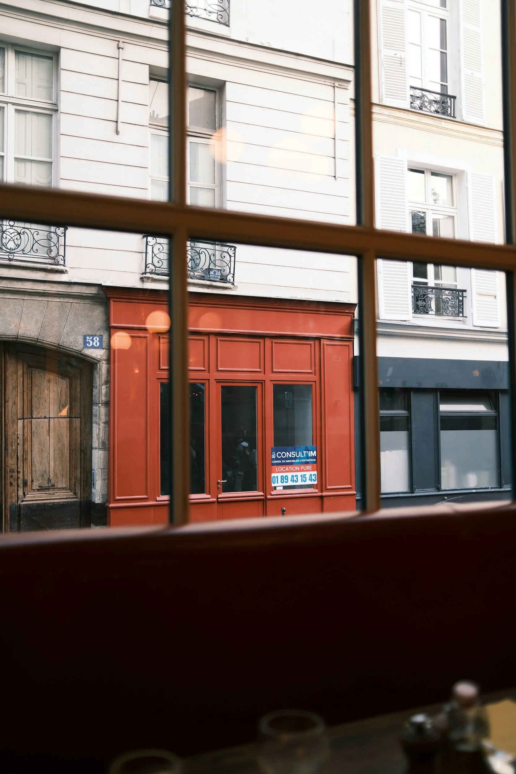 View of a city street seen through a window, featuring a red storefront with a sign and the number 58, and white apartment buildings with balconies and window shutters in the background.