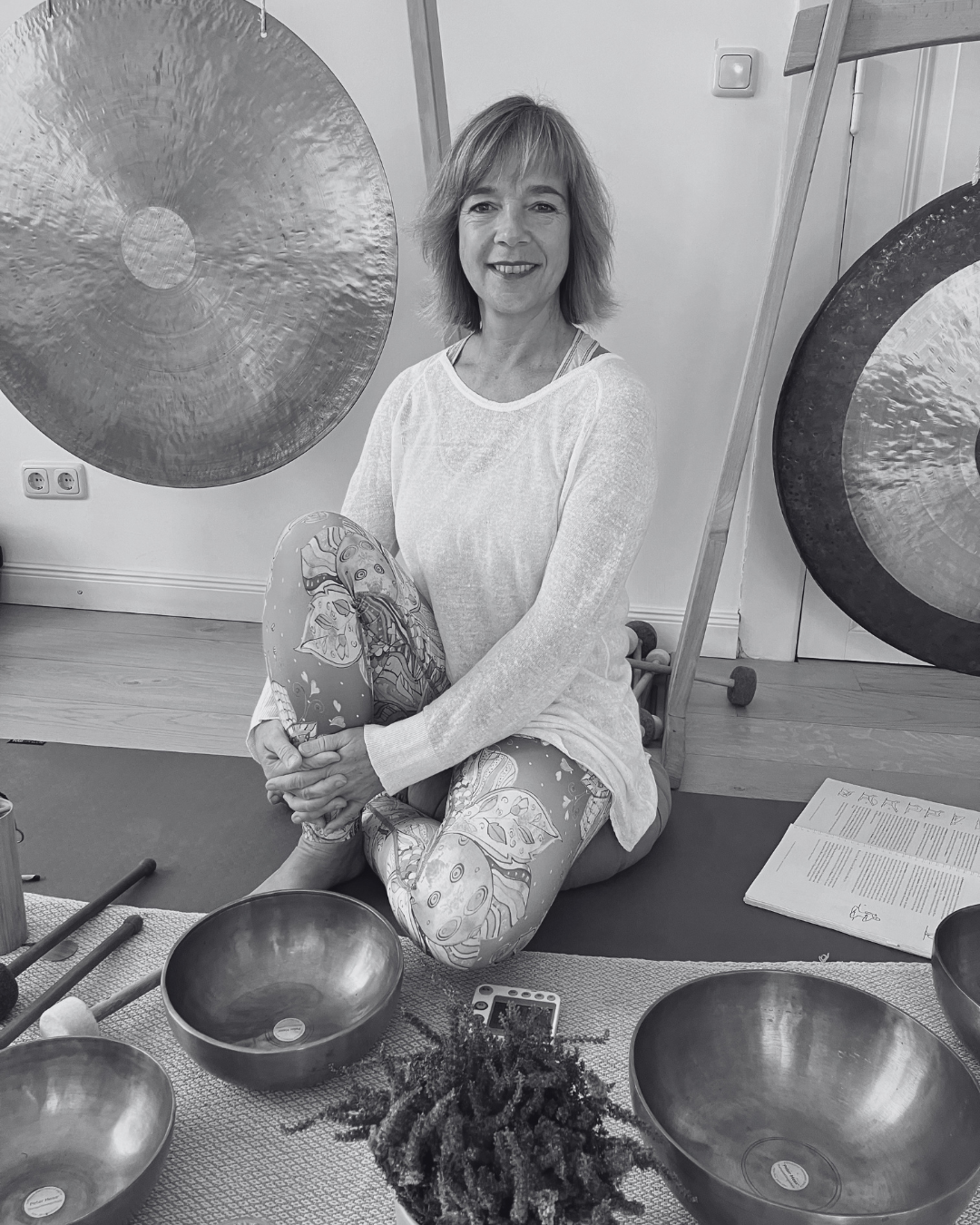 Frau sitzt entspannt zwischen Klangschalen und großen Gongs in einem hellen Studio – Schwarz-Weiß-Foto