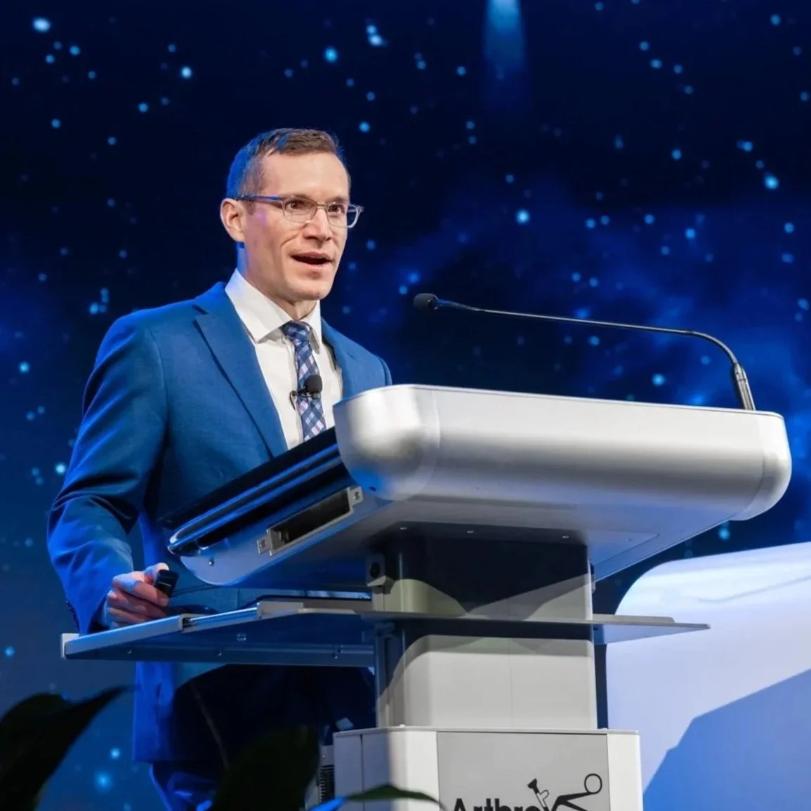 Dr. Derman in a blue suit and glasses standing at a podium giving a speech, with a starry background.