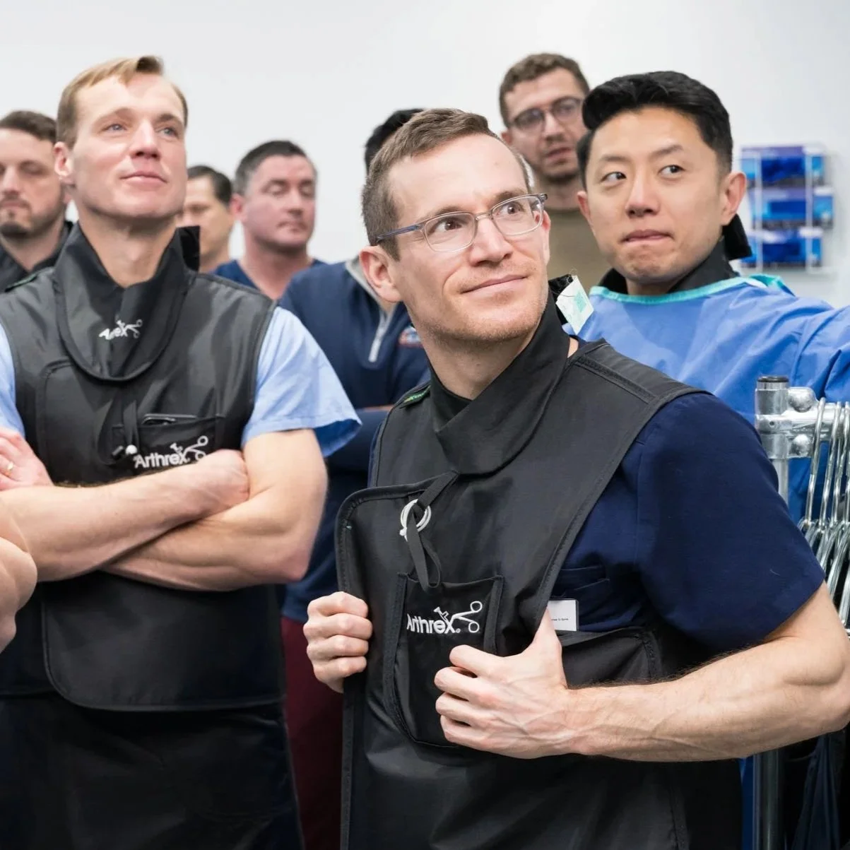 Group of astronauts in training or briefing, with one smiling and adjusting his vest, surrounded by other crew members in a laboratory or training facility.