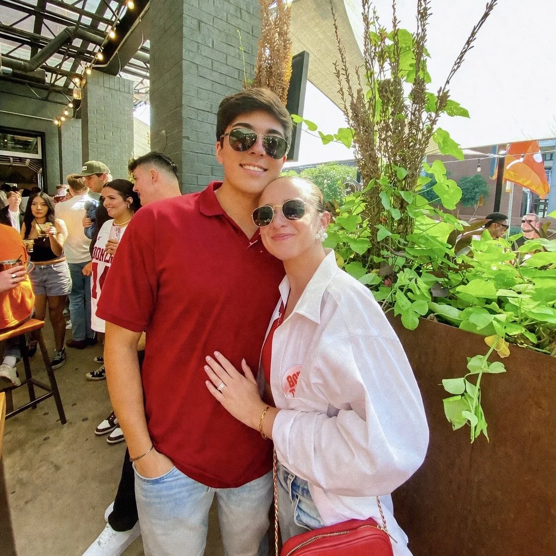 A woman (Claire) and man (her boyfriend) wearing sunglasses, smiling for the camera at a crowded outdoor event. The woman has her hand on the man's chest, and they are standing beside a large green leafy plant.