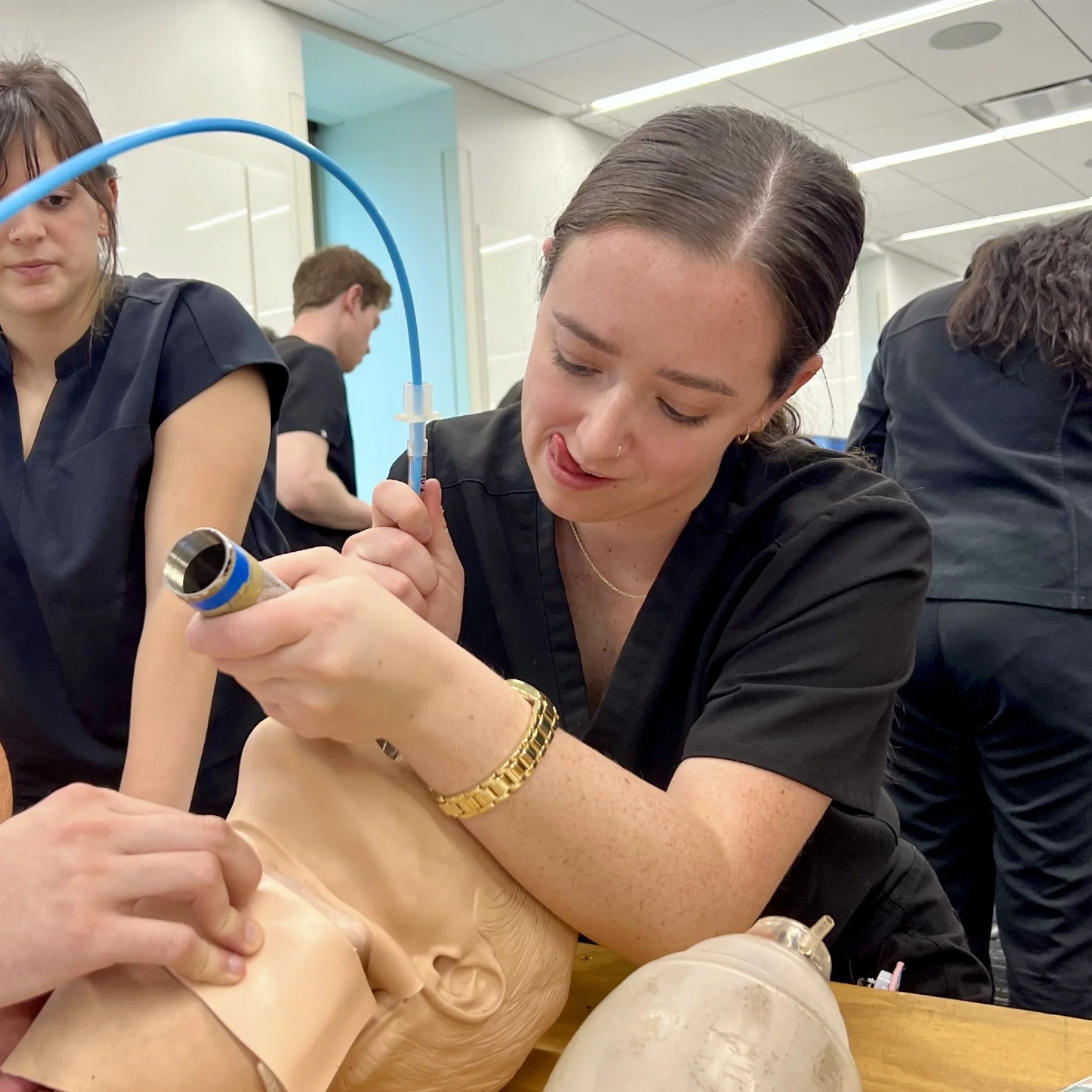 Claire performing a medical procedure on a mannequin. She is wearing dark scrubs and surrounding by others in a training environment.