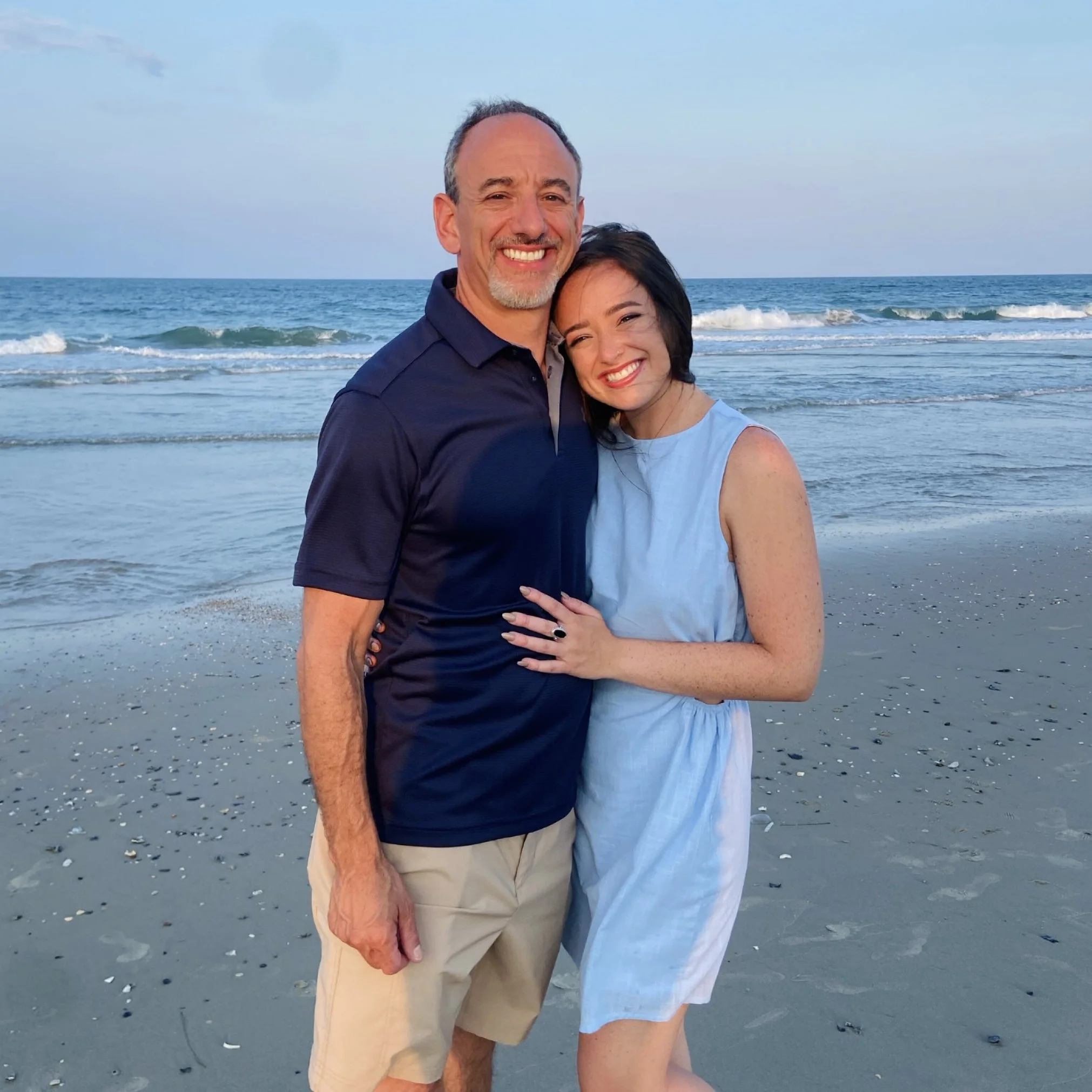 Claire and her father hugging on a beach, smiling at the camera. Waves and the ocean in the background during sunset.
