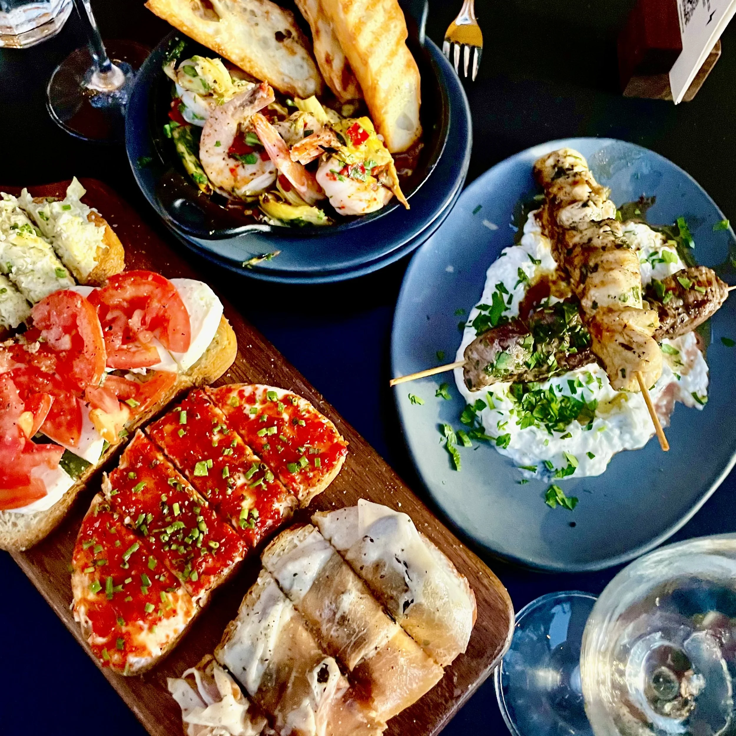 Assorted appetizers on a wooden serving board, including tomato slices, cream cheese with herbs, and various finger foods, with glasses of water and condiments in the background.