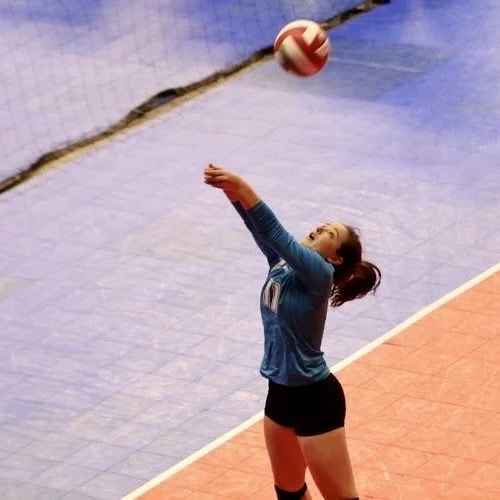 Claire playing in a volleyball match in high school. She is blue wearing a blue jersey and black shorts setting a volleyball during a game inside a gym.
