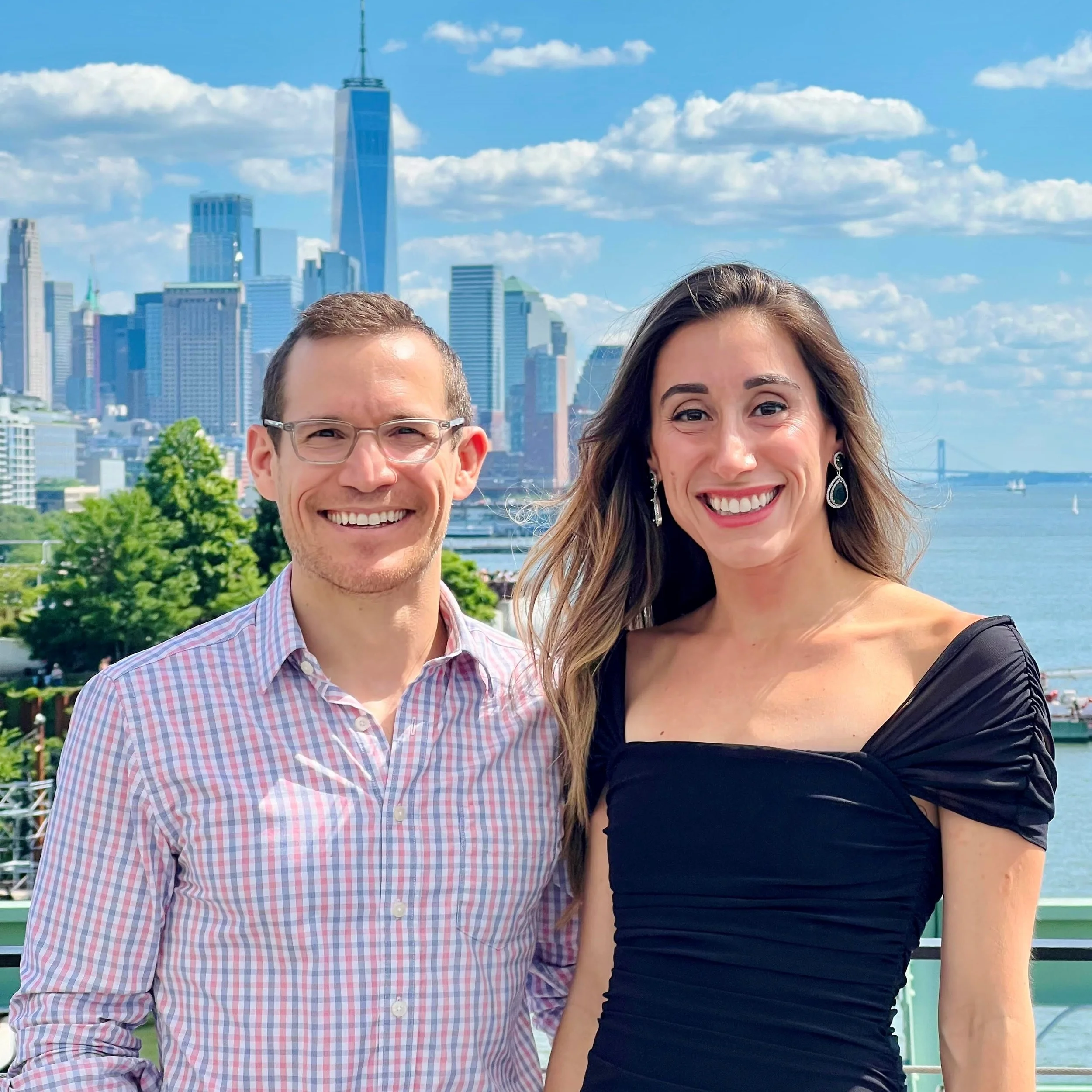A smiling Dr. Derman and his wife standing outdoors with the New York City skyline and water in the background on a sunny day.