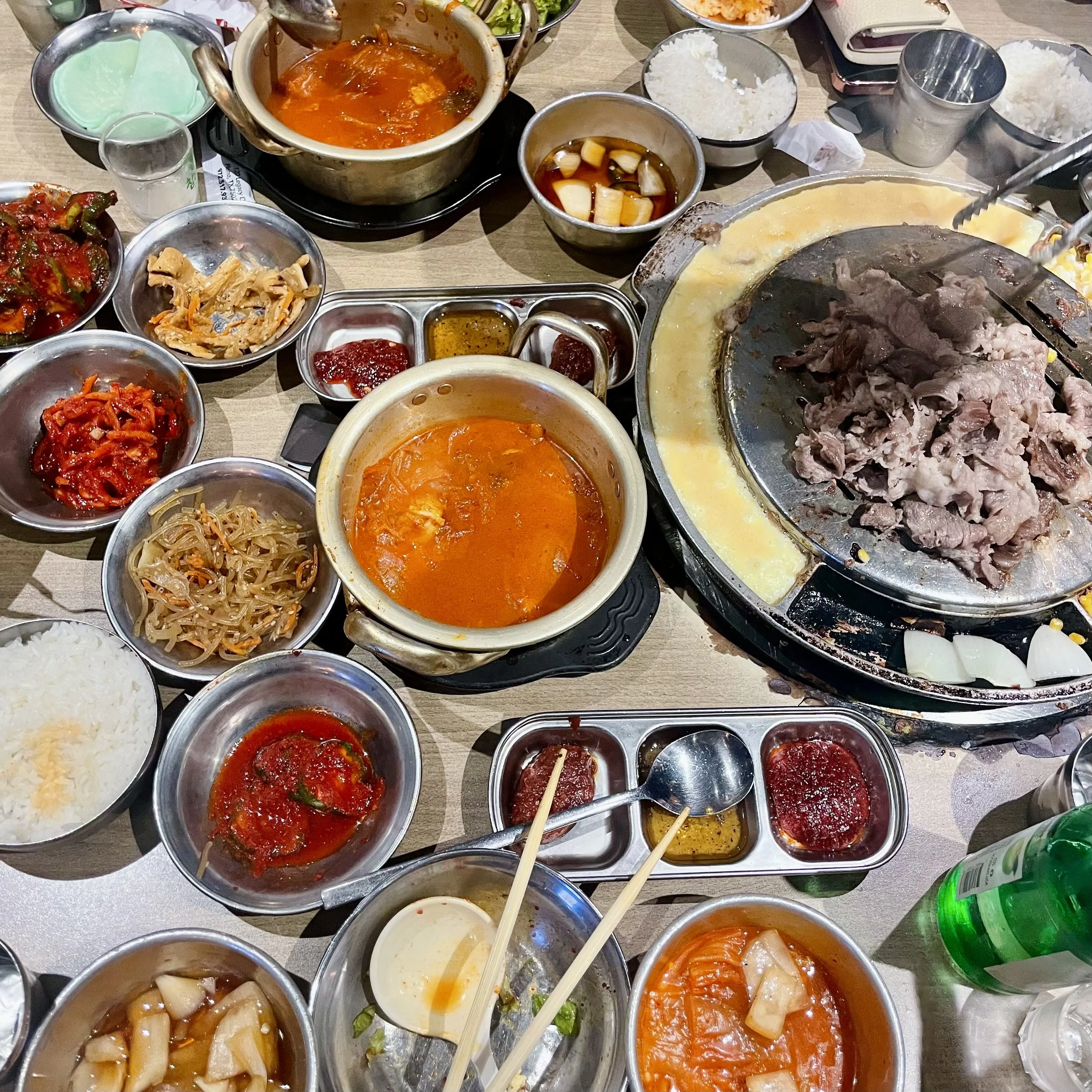 A table filled with various Korean dishes, including bowls of rice, kimchi, dipping sauces, and multiple hot pots with stews and cooked meats.