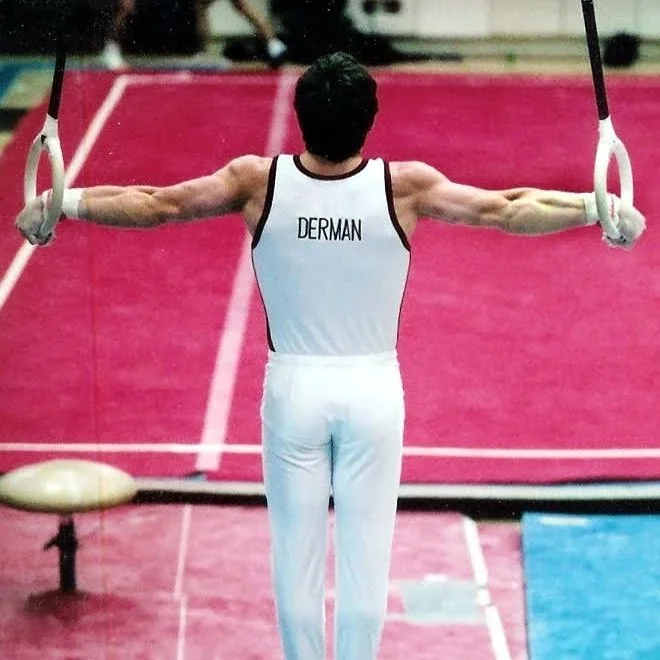 Dr. Derman in a white gymnastics uniform performing an iron cross on the rings at a gymnastics meet. His back is to the camera, and the name 'DERMAN' is printed the back of his uniform.