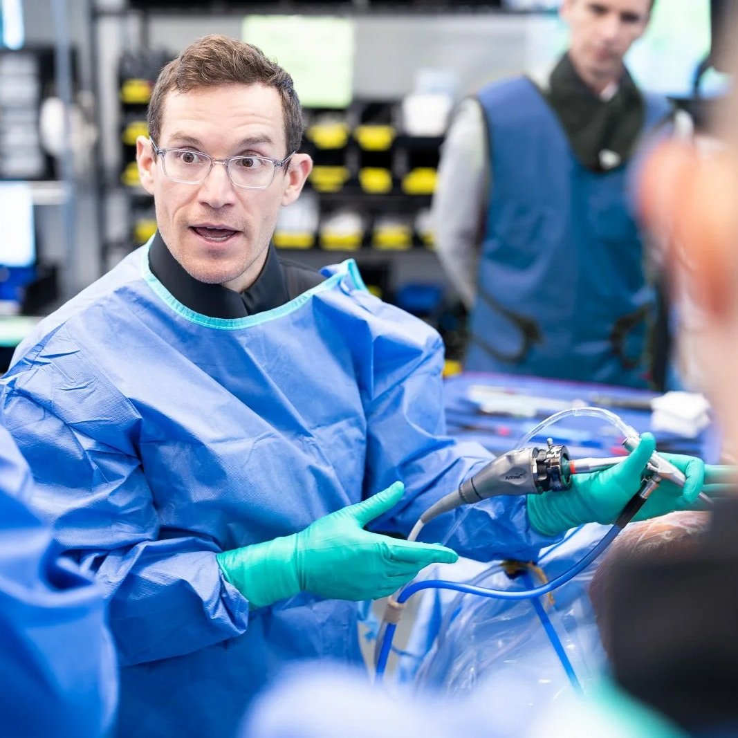Dr. Derman in a surgical gown and gloves scrubs demonstrating and explaining an endoscopic procedure to a group, holding and endoscope, in a lab setting.