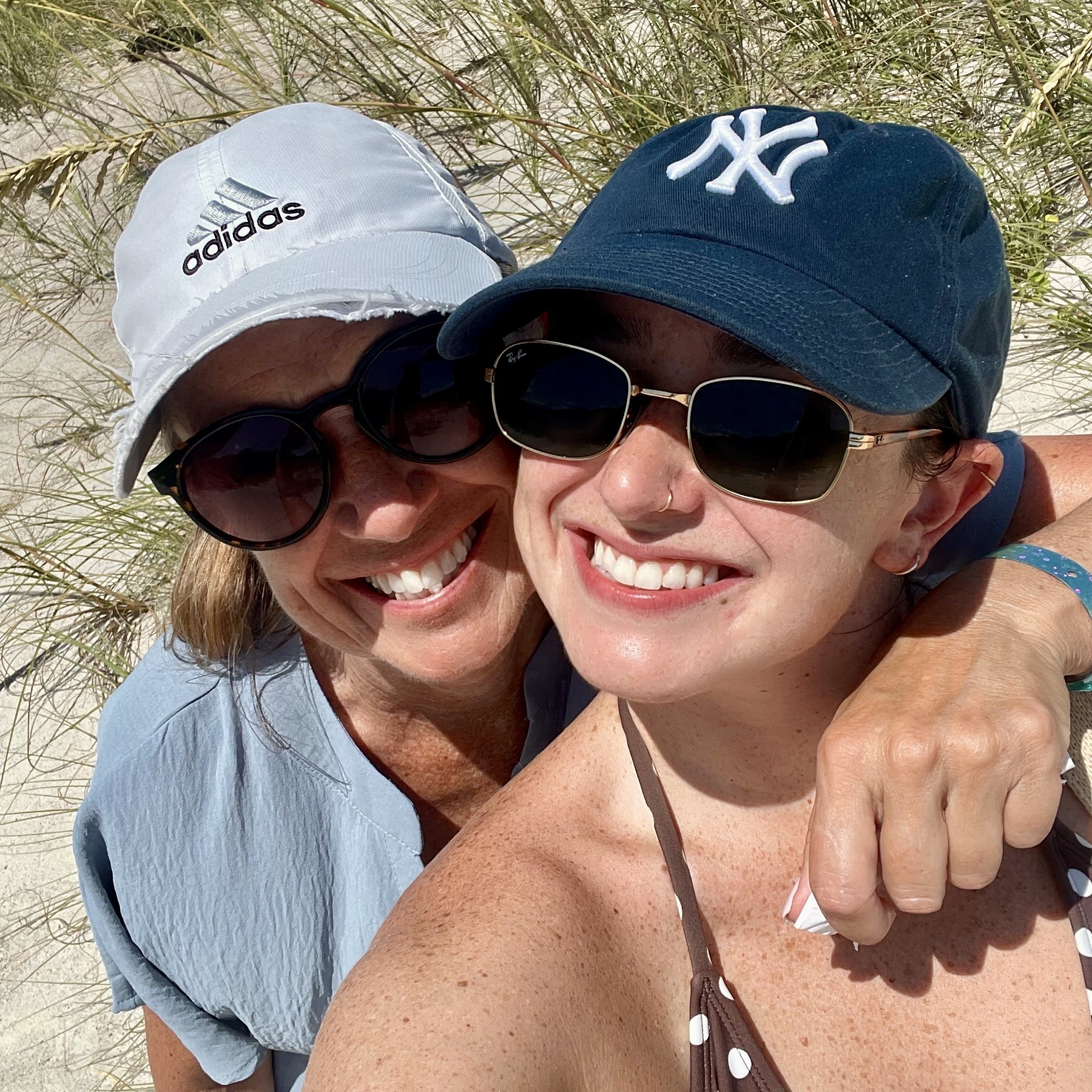 Close-up of Claire and her mother smiling at the beach. Claire is wearing a blue New York Yankees cap and sunglasses, and her mom is wearing a white Adidas cap and sunglasses, with beach grass and sand in the background.