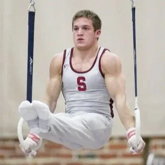 Male gymnast performing on rings with focused expression, wearing a white athletic uniform with red trim and an 'S' logo.