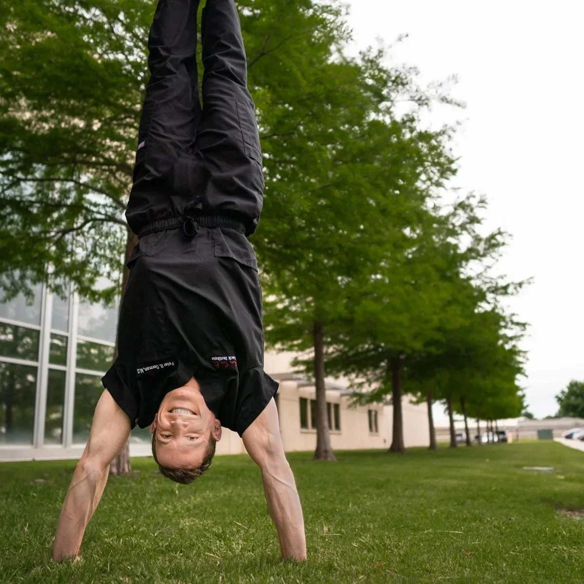 Dr. Derman performing a handstand outdoors on grass, smiling with trees and a building in the background.
