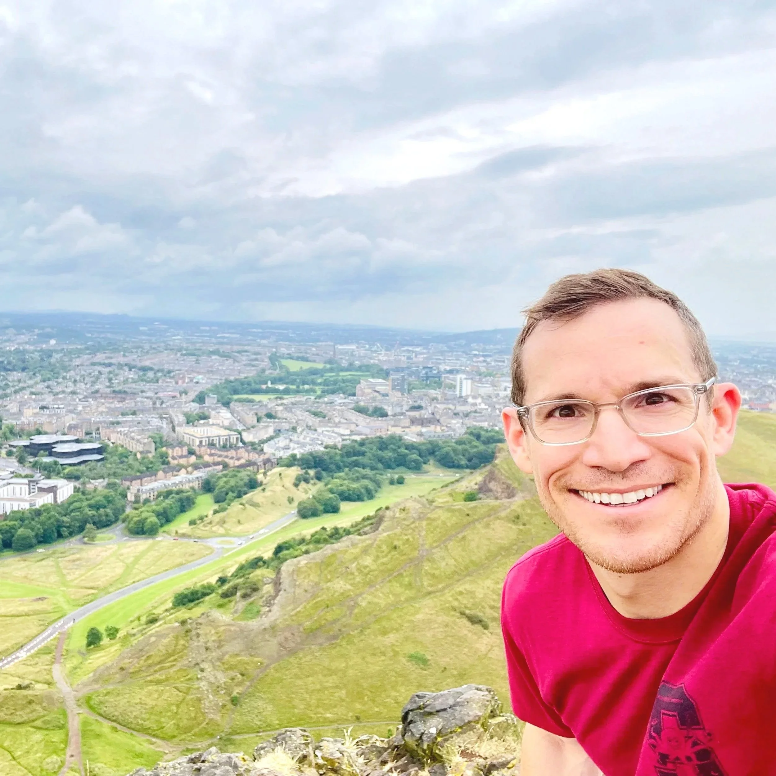 Dr. Derman smiling outdoors with a city and green landscape in the background, wearing glasses and a red T-shirt, with the top of their head and part of the face visible.