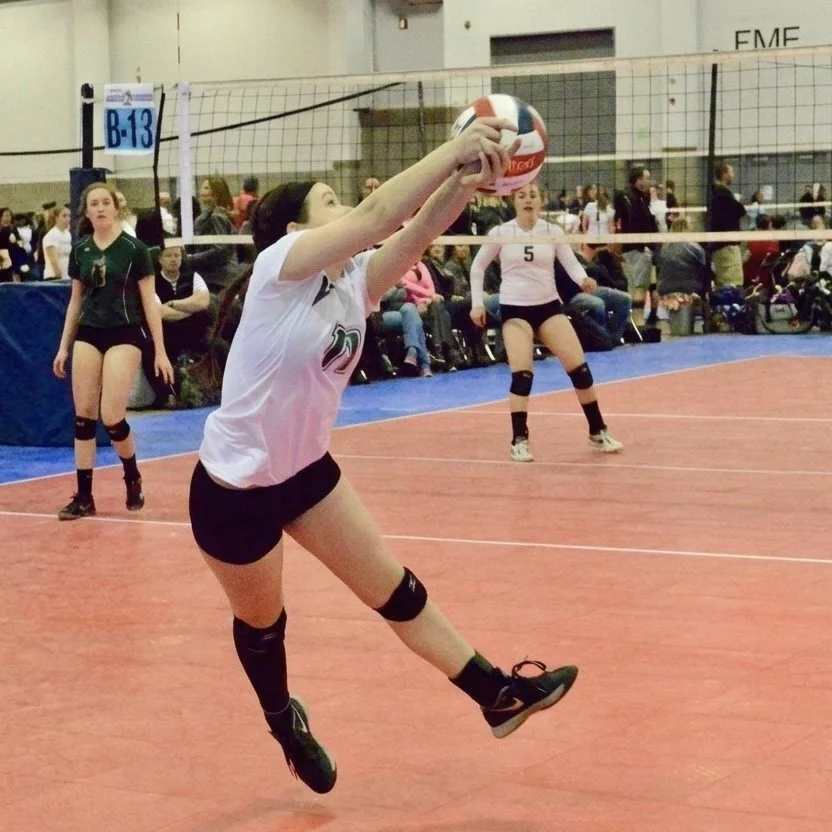 Claire in a white jersey and black shorts jumps to hit a volleyball in an indoor court, with other players and spectators in the background.