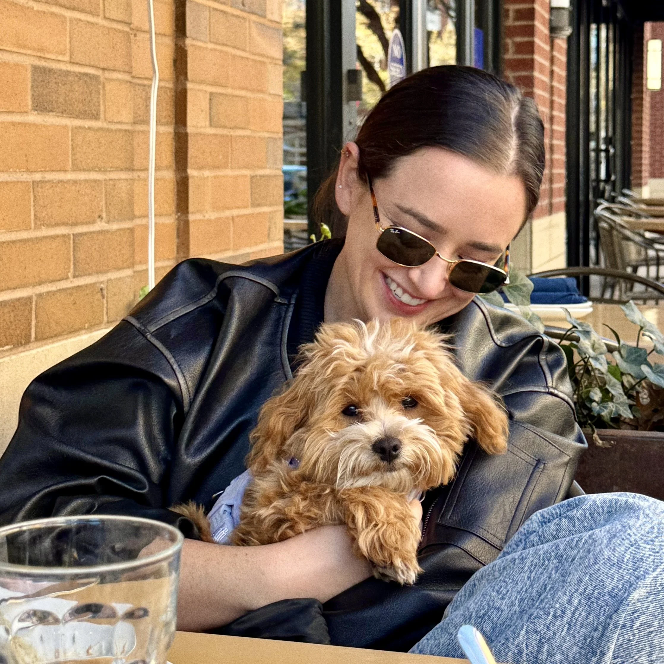 Claire, a young woman wearing sunglasses and a leather jacket, sitting outside at a restaurant holding her fluffy, cute tan dog.