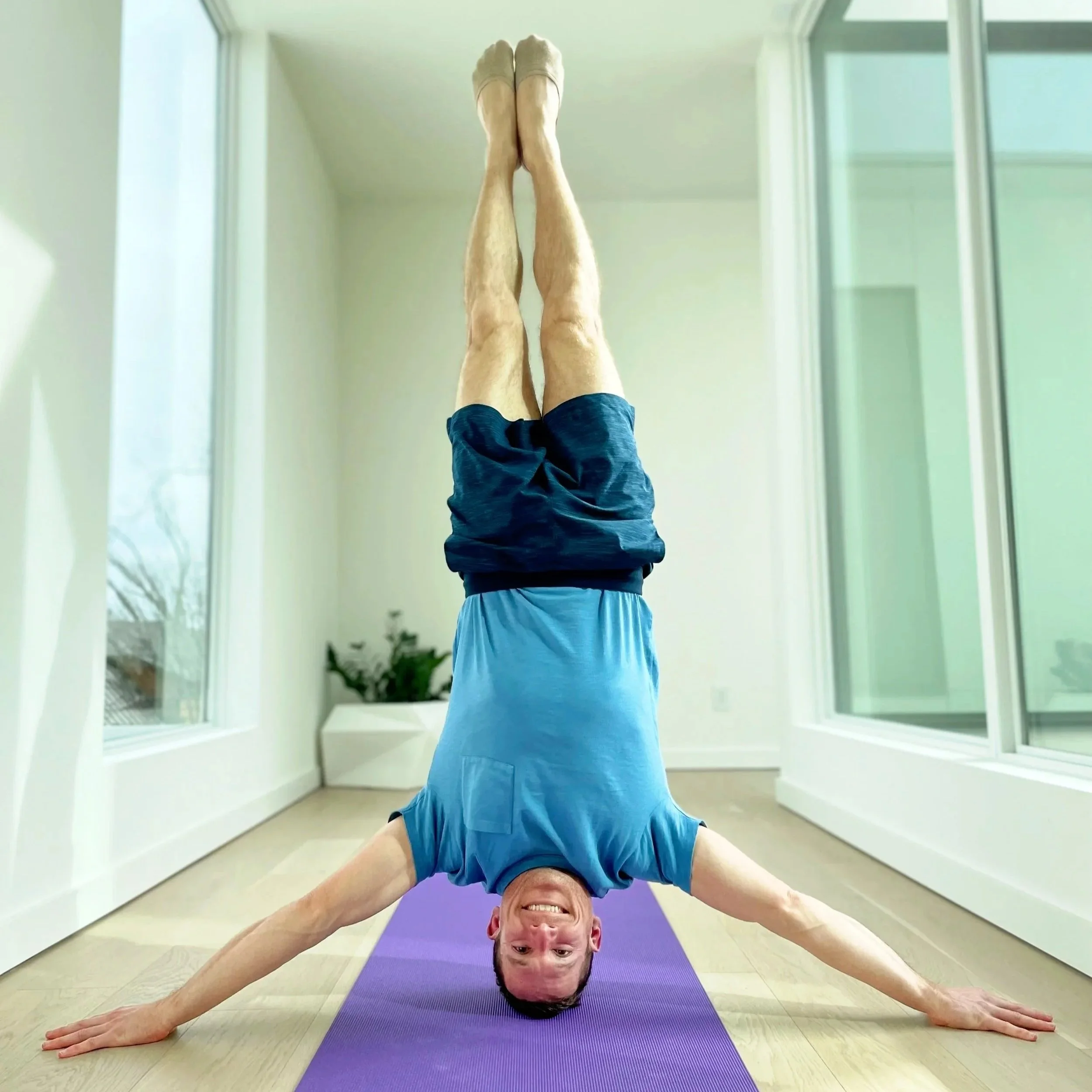 Dr. Derman performing a yoga pose on a purple mat in a bright room with large windows, wearing a gray shirt and blue shorts, with his body upside down and hands on the floor.