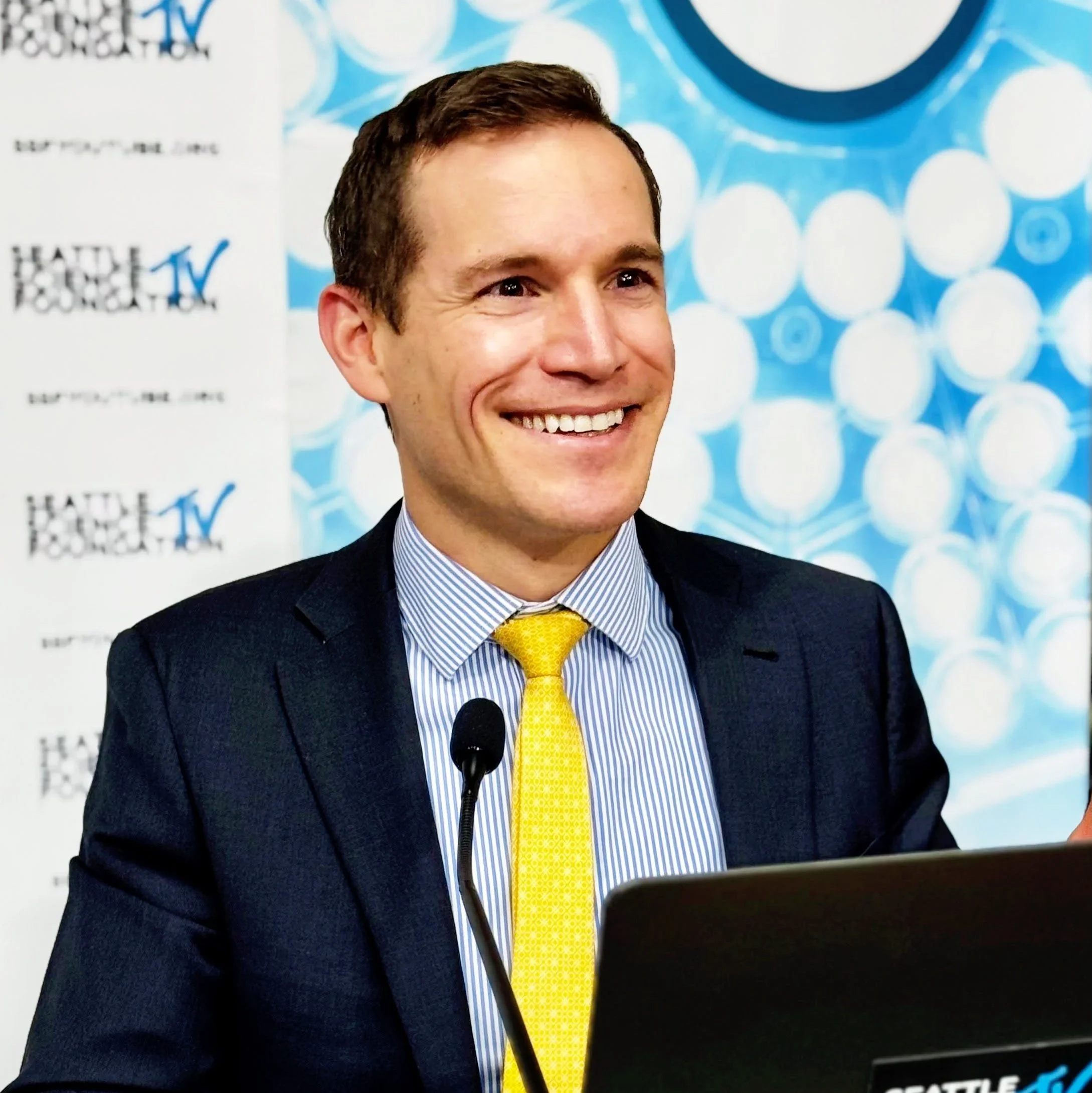 Close-up of a smiling Dr. Derman in a dark suit with a yellow tie and light blue striped shirt, standing at a podium in front of a blue and white background.