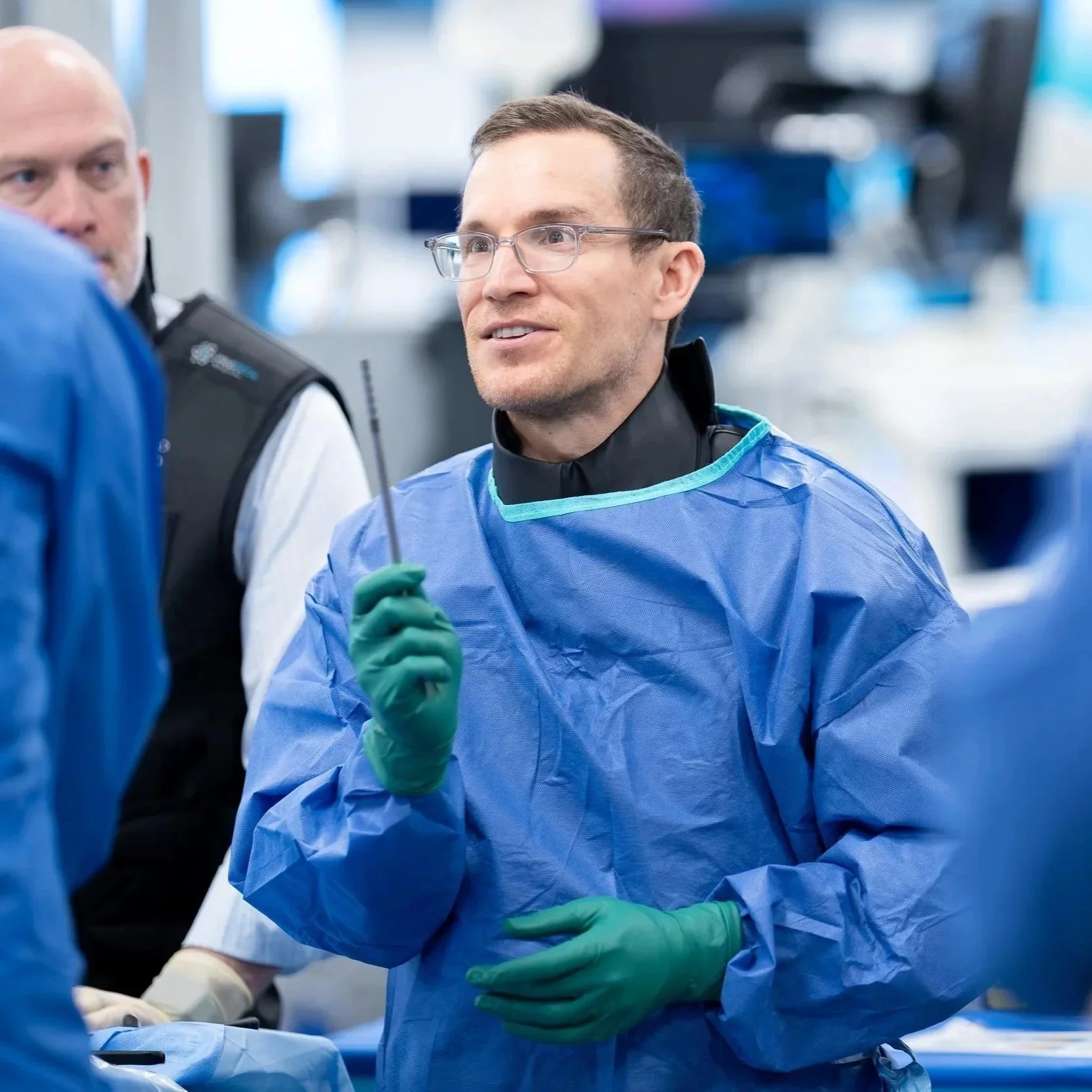 Dr. Derman teaching other surgeons. He is wearing glasses and a blue medical gown holding a thin endoscopic instrument, in a lab setting with another person and medical equipment in the background.