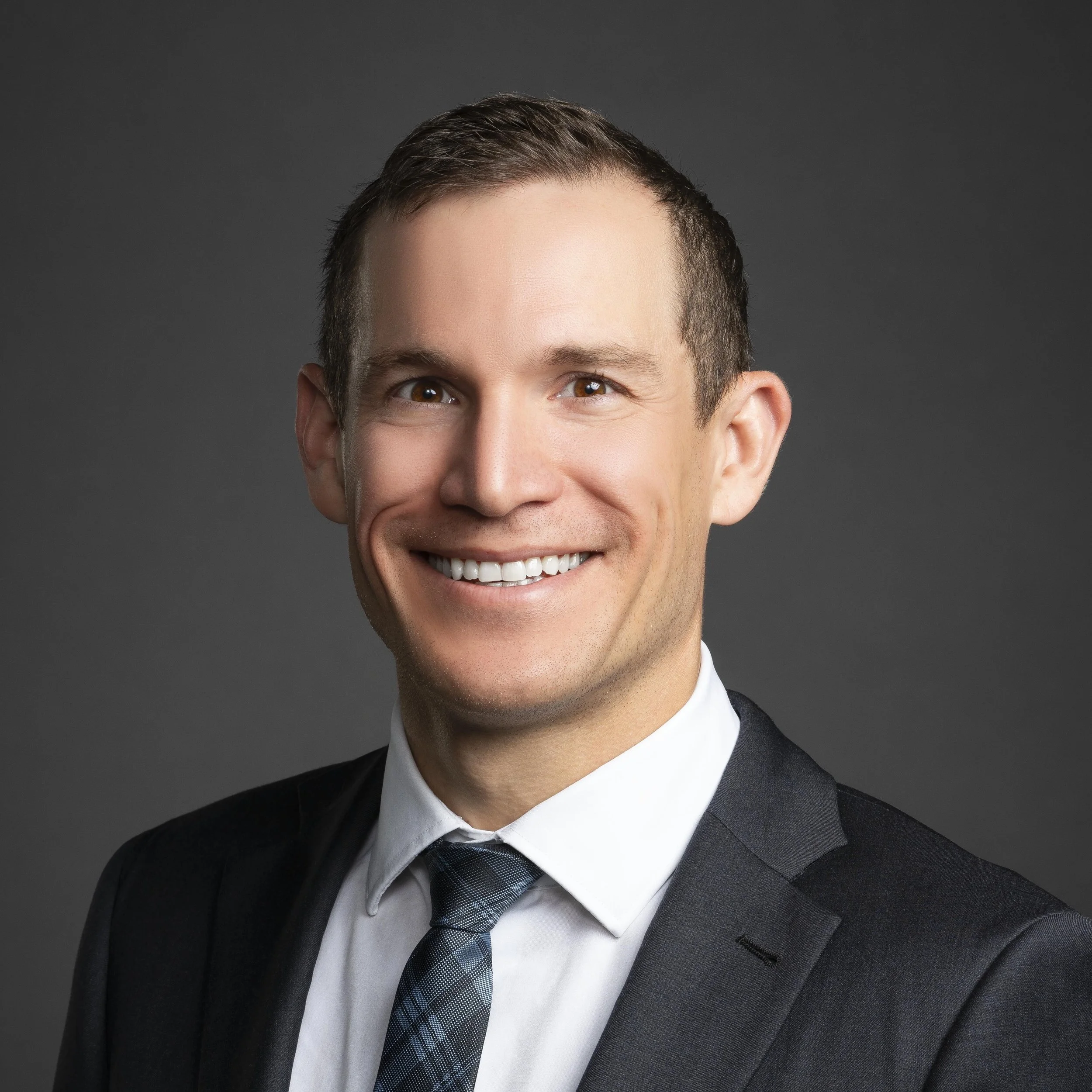 Close-up of Dr. Derman, smiling and dressed in a black suit, white shirt, and dark plaid tie, against a dark gray background.