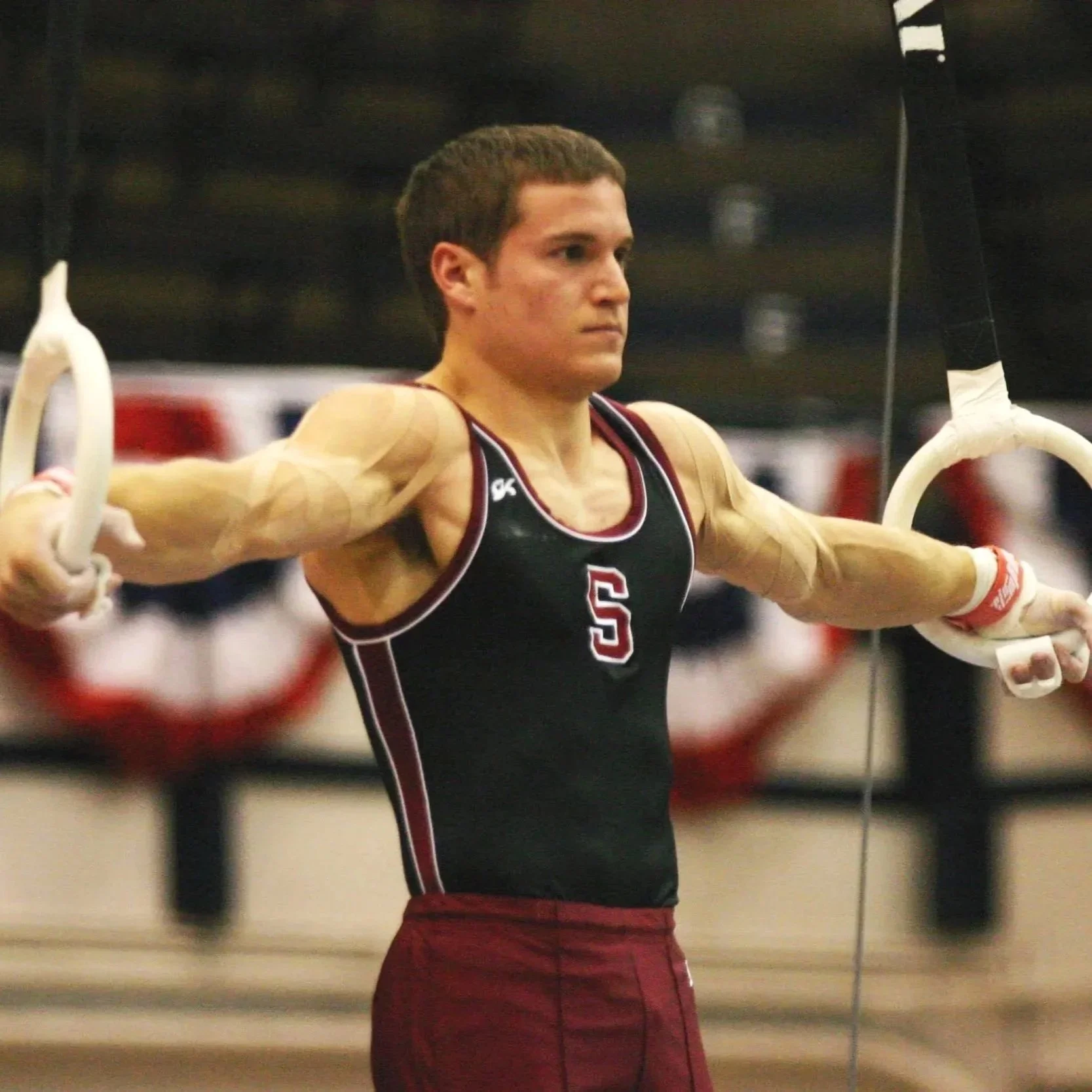 Dr. Derman performing an iron cross on the rings at a gymnastics meet in college while wearing a Stanford gymnastics uniform.