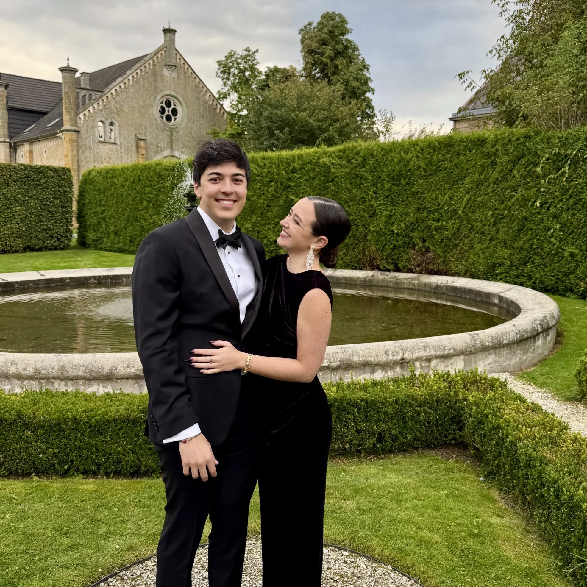 A happy young man (Claire's boyfriend) in a tuxedo and a woman (Claire) in a black dress smiling and looking at each other outdoors near a stone fountain with lush greenery and a church in the background.
