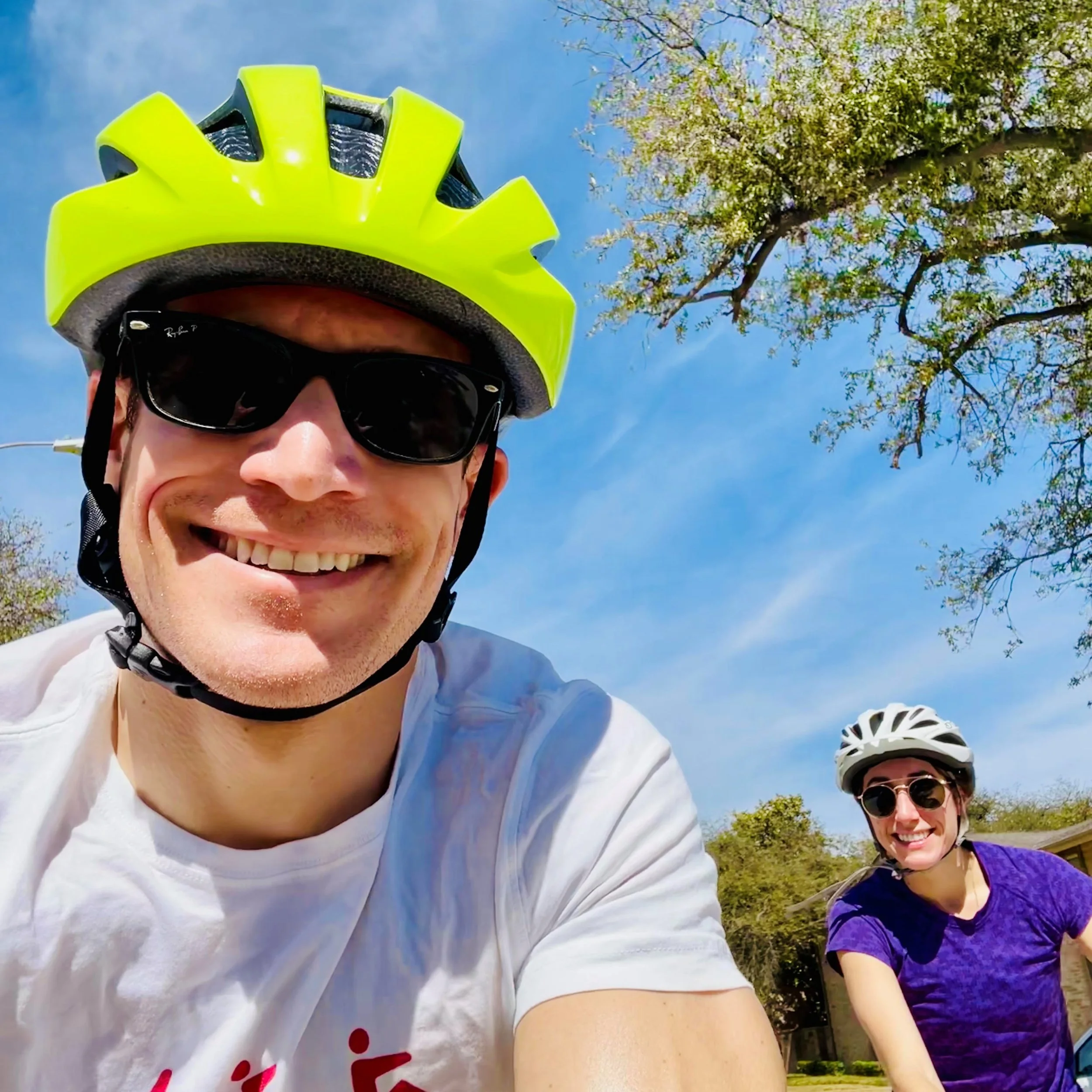 Close-up of Dr. Derman and his wife outdoors on bicycles, wearing helmets and sunglasses, with trees and blue sky in the background.