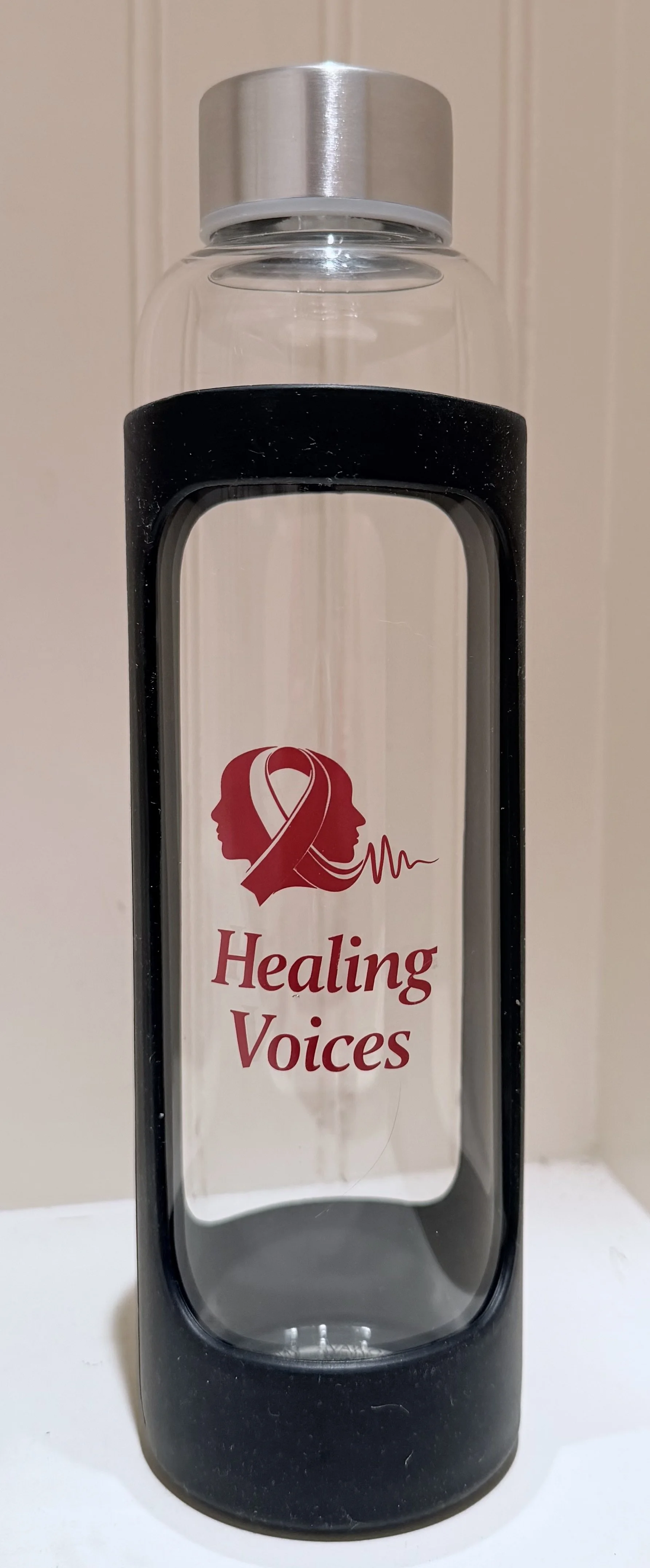 A clear water bottle with a black rubber sleeve and a silver lid. The bottle has a red logo of two faces with a ribbon and a heartbeat line, and the words "Healing Voices" underneath.