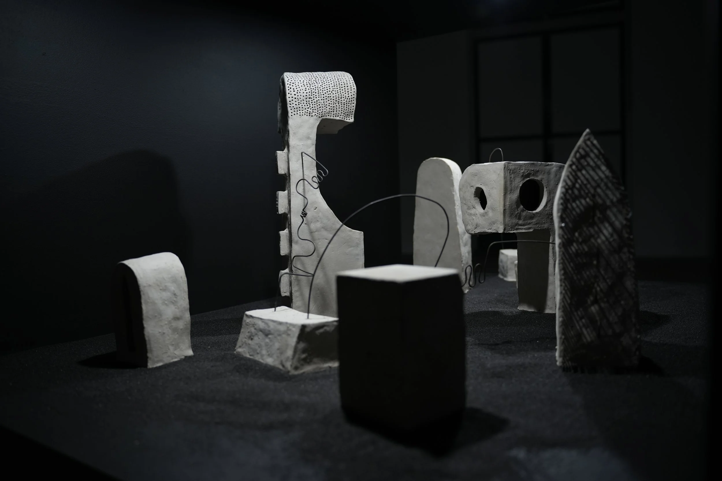 Zu sehen ist die landschaftliche Installation verschiedener, durch Julian Schomäker, gefertigter, roh belassener Tonskulpturen. Die Skulpturen sind durch Drähte miteinander verbunden und sind auf schwarzem Sand in einem Kasten installiert.